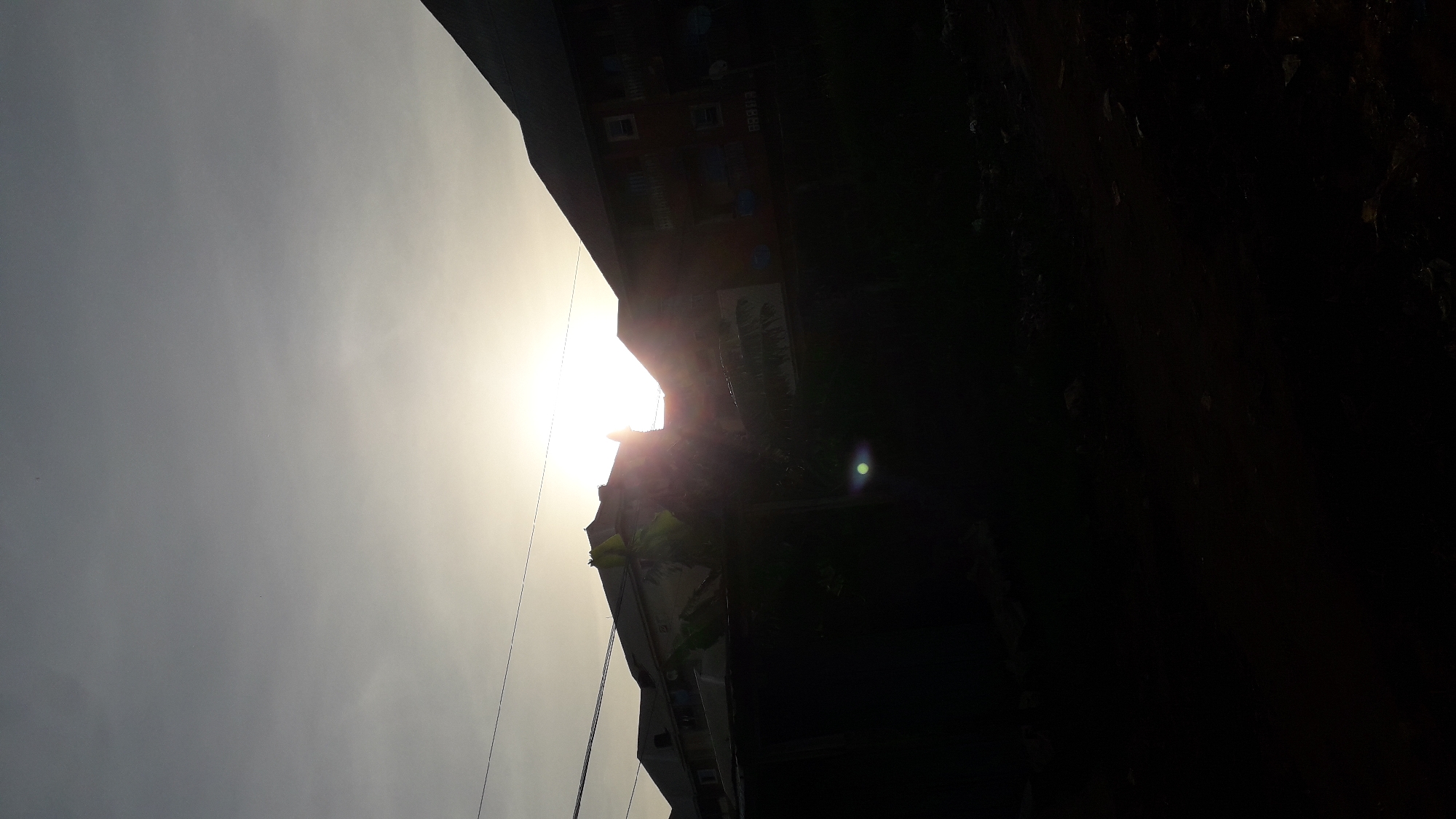 The image depicts an urban scene in Nkpor, Nigeria, with a building silhouetted against a bright sky dominated by the sun. Power lines are visible against the sky. The right side of the image is largely obscured by dark shadows. The architecture suggests a multi-story residential or commercial building.