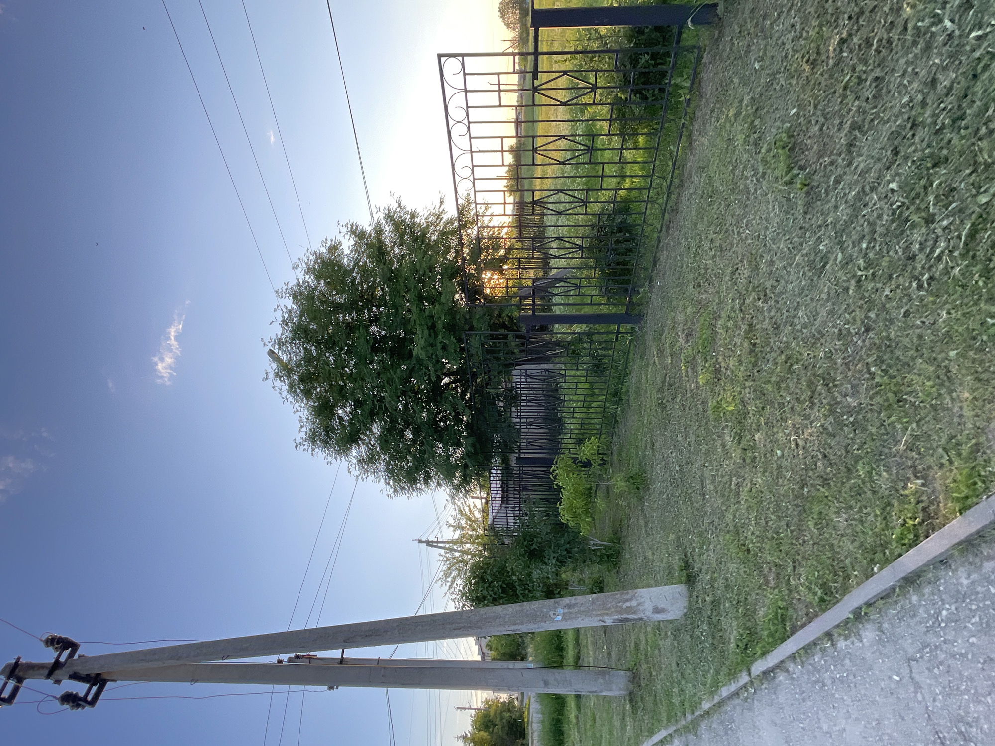 The image depicts a scene in the city of Krasnye Baki, Russia, on a clear day with a bright blue sky. In the foreground, a concrete sidewalk runs along the left side of the frame, bordered by a strip of mown grass. Two concrete utility poles stand tall on the right of the sidewalk, supporting electrical wires that stretch across the image. Beyond the grass, a black metal fence with decorative elements encloses a yard. A lush green tree stands within the fenced area, its branches reaching towards the sky, and sunlight filters through its leaves. The fence continues to the right, revealing a grassy field or yard beyond, with more trees visible in the distance under the setting sun. The overall impression is a quiet, residential area in the late afternoon or early evening.