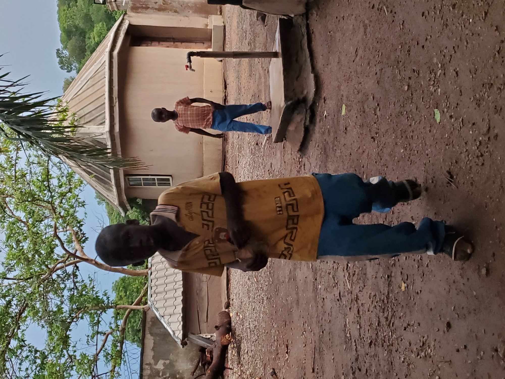Two boys are in a dirt yard in Mararaba, Nigeria. The foreground boy wears a yellow shirt and blue pants. The background boy wears a plaid shirt and blue pants and stands near a metal water tap. Buildings and trees surround the yard.