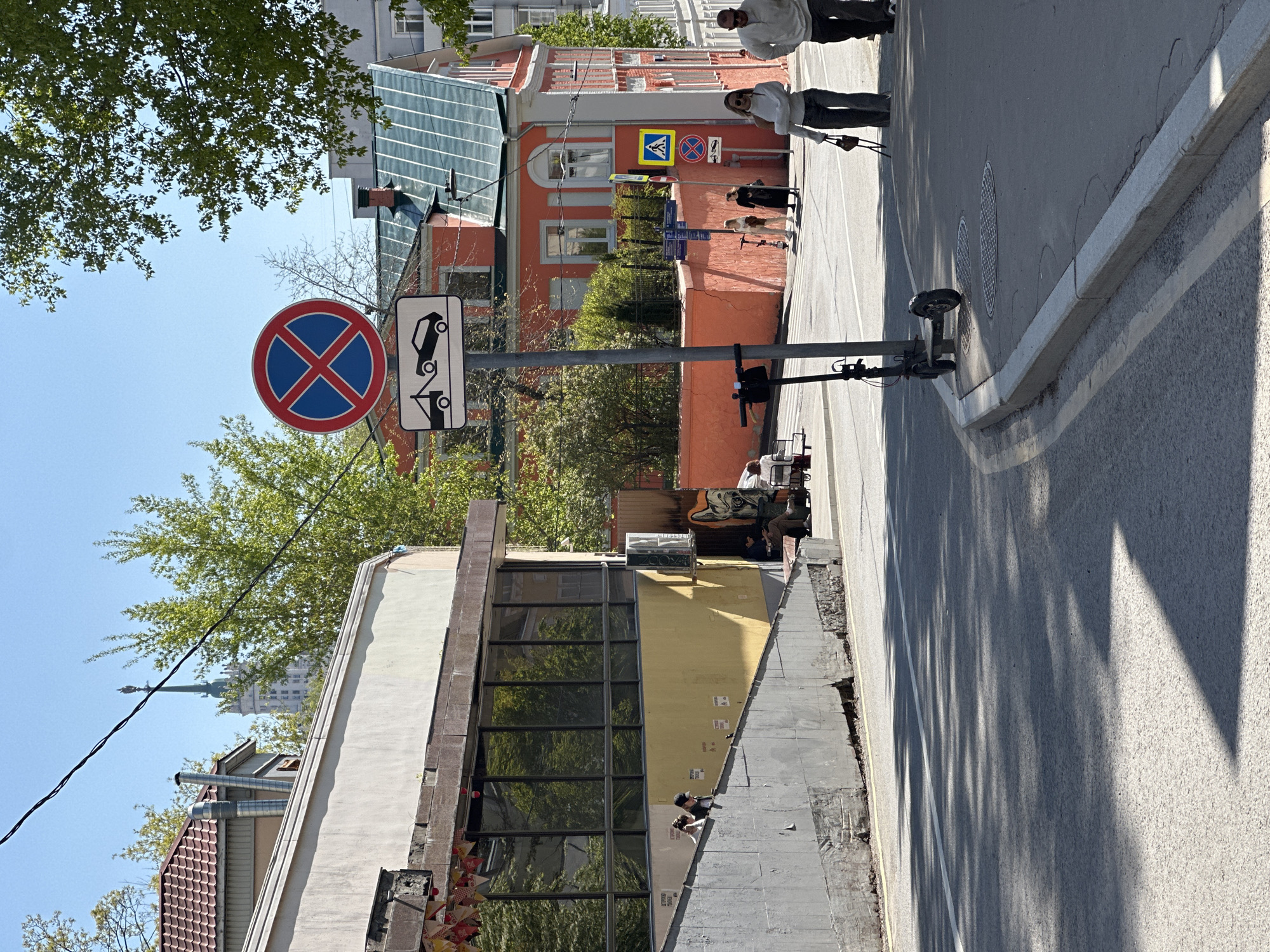 The image captures a street scene in Moscow, Russia, featuring a "No Stopping" sign (circular red and blue X on white background) accompanied by a sign indicating that vehicles will be towed. The background showcases a multi-story building, appearing light peach in color with several windows and a pedestrian crossing traffic sign. People are walking on the sidewalk, and the street is lined with a curb. Above the buildings, a blue sky is visible, with a building appearing to be one of the "Seven Sisters," Stalinist Skyscrapers, in the far distance. A scooter is parked along the curb. A crew appears to be working on a building on the right side.