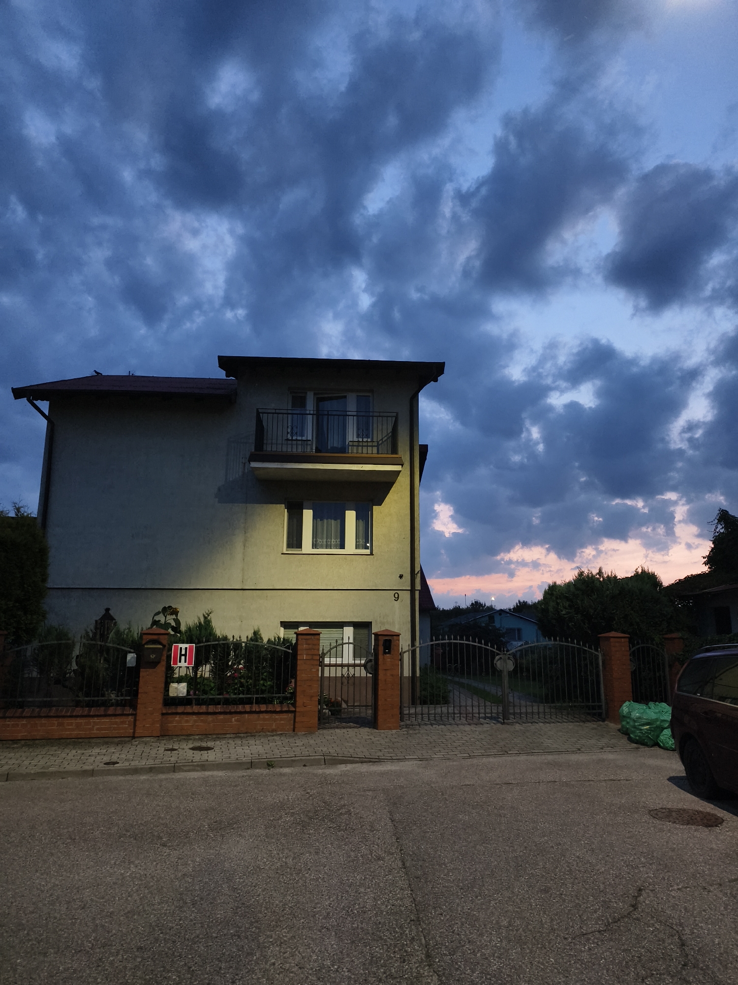 This is a low-angle, exterior, medium shot captured at dusk of a two-story building with a balcony, situated in Tczew, Poland. The building is light yellow and features a red-tiled roof. A black metal fence with brick pillars encloses the property. On the fence, a red and white sign with the letter "H" is visible. To the right of the fence, a green bag of trash is placed near a dark-colored vehicle. The sky above is filled with dark, dramatic clouds. The scene appears to be a residential or commercial property on a street with paved asphalt and cobblestone sidewalks.