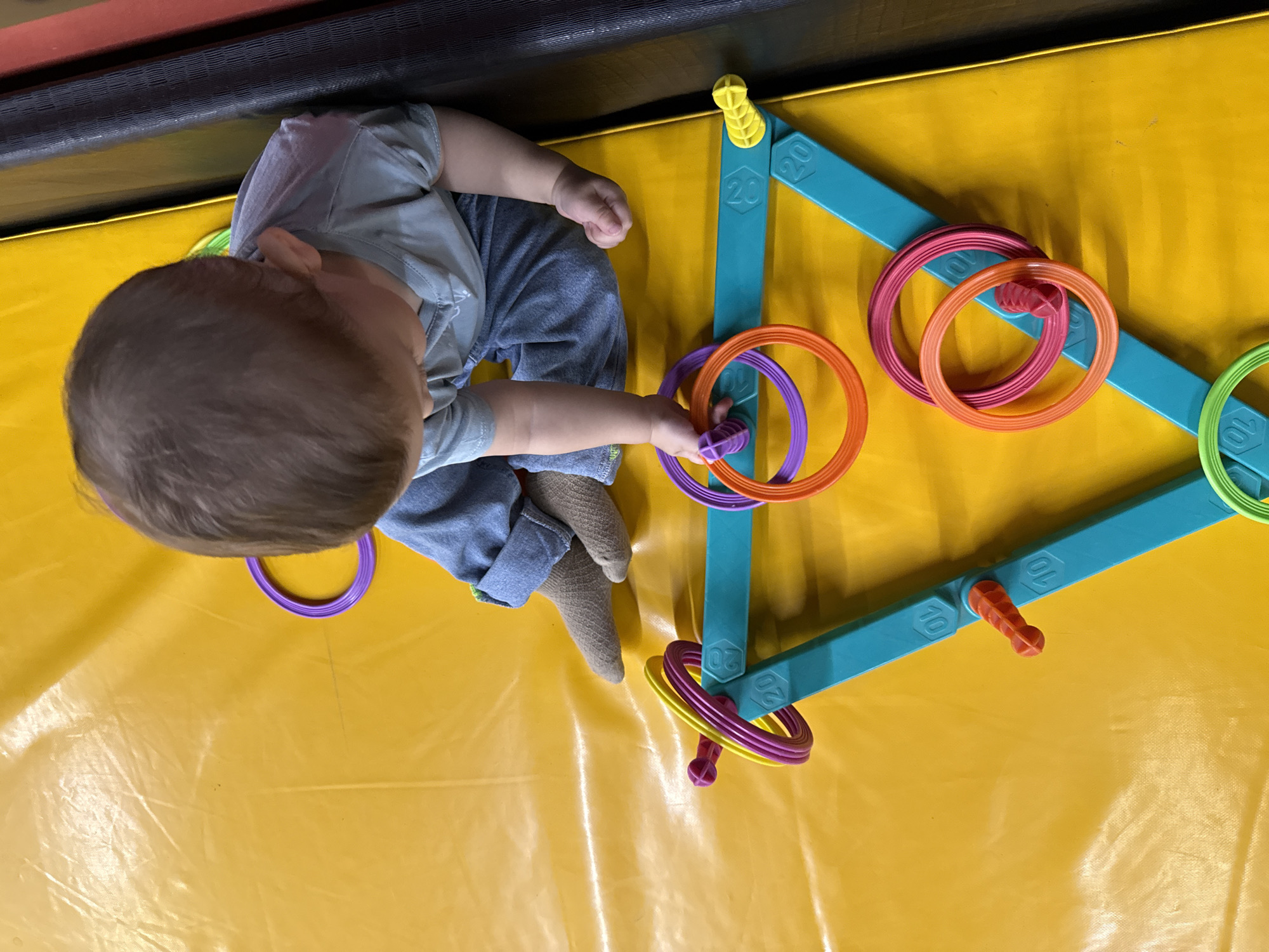 An overhead, medium shot captures a young child sitting on a bright yellow, padded surface, possibly a gym mat, in a brightly lit indoor space. The child, with brown hair and wearing a light blue t-shirt and blue jeans, is looking down at a colorful ring toss toy. The toy is turquoise and shaped like a triangle with pegs and numbered markings. Several rings in shades of purple, orange, pink, and yellow are scattered around the toy. The child's hands are engaged with the toy, and their legs are crossed. The background shows the edge of the yellow mat transitioning into dark and red vertical padding, suggesting an indoor play area or gymnasium. The location is not specified in the image.