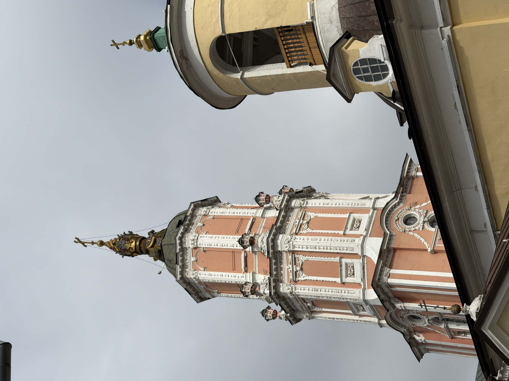 The image shows two church towers against a gray sky. The lower tower is primarily salmon-colored with white trim, elaborate ornamentation, and a golden spire. The second tower is yellow with a rounded top section featuring an arched opening. A building structure is partially visible in the foreground, with a beige wall on the right. The photo appears to have been taken at a low angle, looking up at the buildings. The structures may be located in the city of Moscow, Russia.