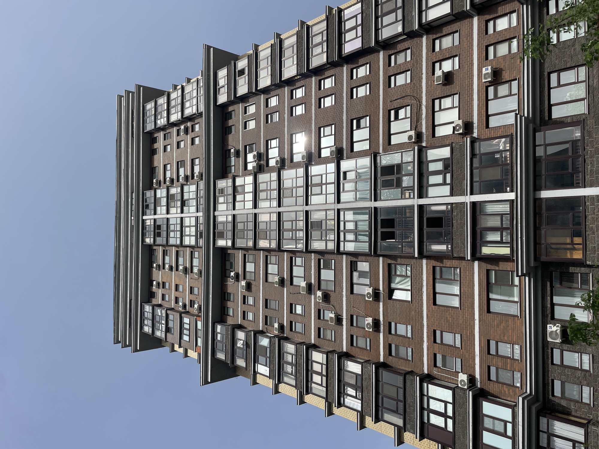 The image depicts a high-rise brick building with multiple windows and air conditioning units, seen from a low angle. The building has a modern design with vertical gray elements running along its length. The sky is clear blue. The location is Kazan, Russia.