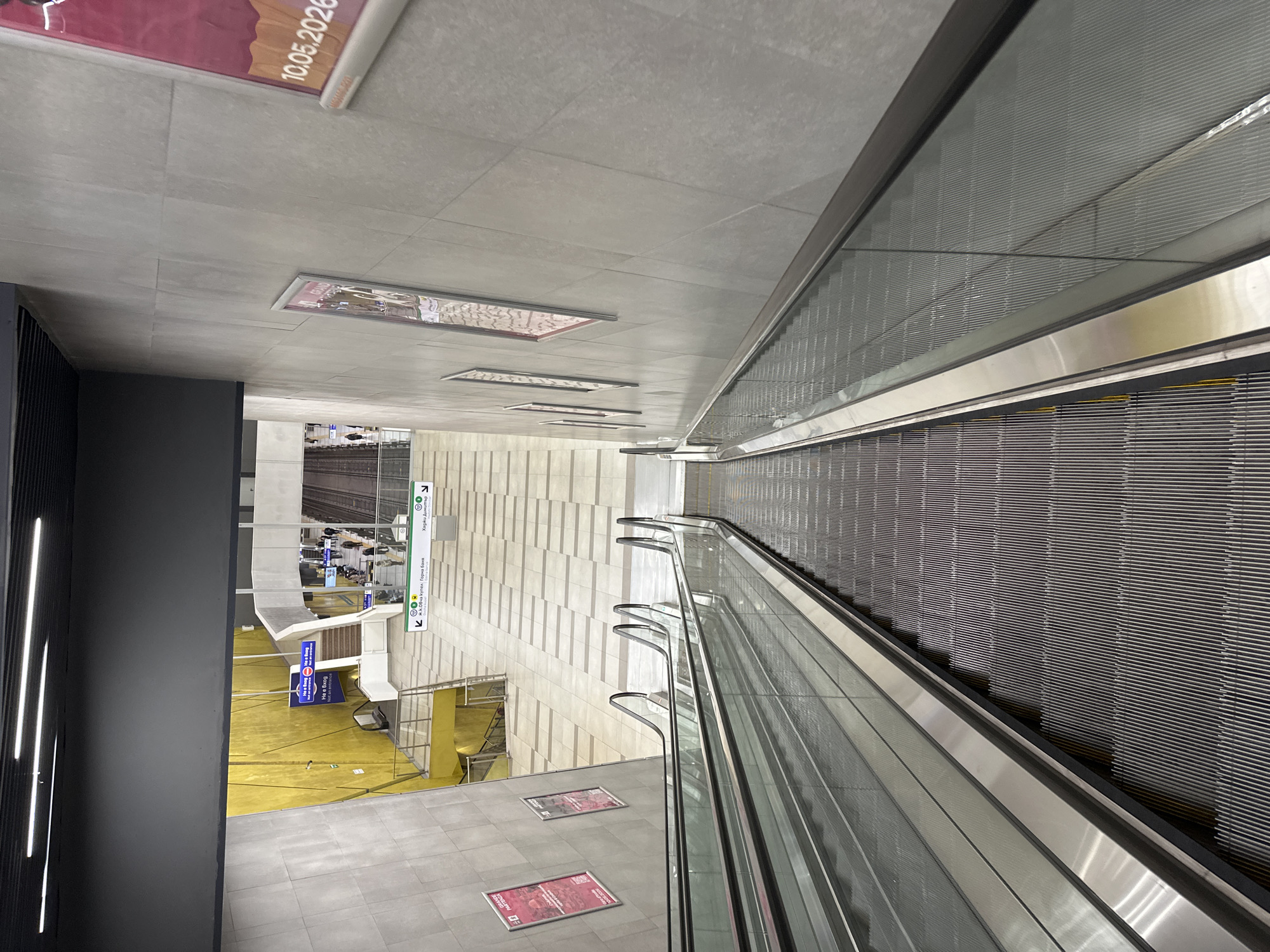 This is an indoor, eye-level shot of an escalator leading down into what appears to be a station in the city of Sofia, Bulgaria. The escalators are silver with glass sides, and the steps are a dark gray color. To the left of the escalator is a gray wall with vertical white lights. The floor is also gray. The wall behind the escalator is cream-colored and has vertical stripes. There is a yellow floor visible below, with signage in Bulgarian. A sign on the wall has the name of a station, "Хаджи Димитър", and the location "К Ж.К.Овча купел Горна баня". There is another sign with the date "10.05.2020" near the ceiling.
