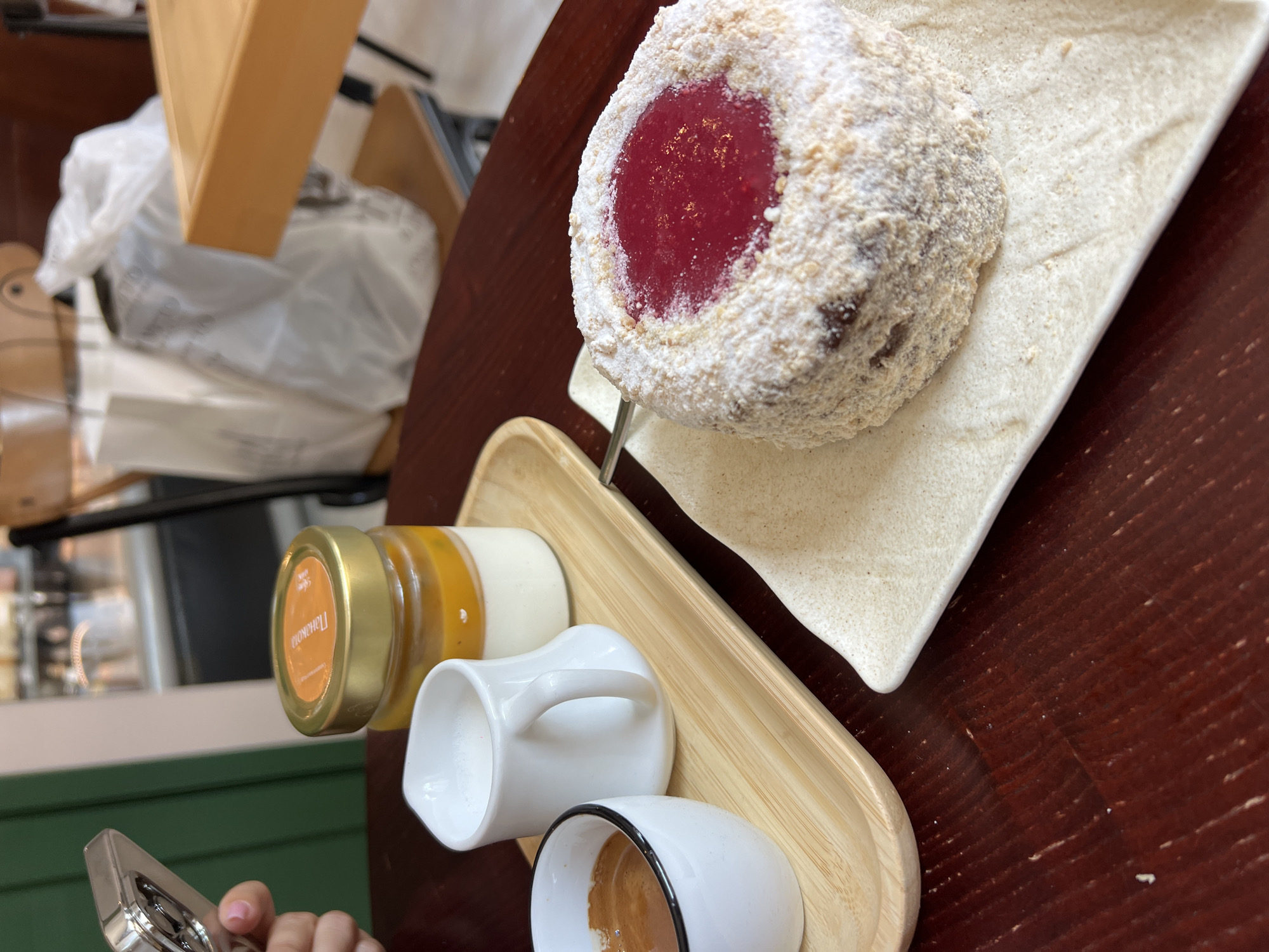 On a dark wooden table, a light-colored square plate holds a small round cake dusted with white powder and sprinkled with crumbs, with a red jam filling in the center. To the left, a wooden tray holds a small white pitcher filled with milk, a white cup containing coffee, and a small glass jar with a golden lid containing two layers of dessert, one yellow and one white. In the background, a white plastic bag sits on a chair next to a wooden table. A person's hand, holding a silver object, is visible in the upper left corner of the frame. The scene appears to be in a cafe or restaurant.