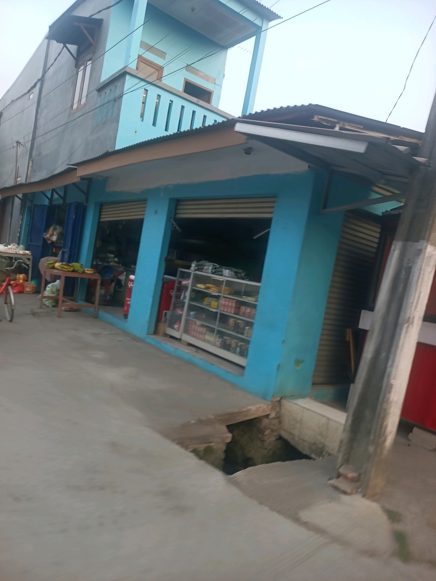 This image depicts a street scene in Telagaasih, Indonesia, featuring a bright blue building that appears to be a small shop or convenience store. The building has large, open storefronts with rolled-down shutters visible inside. To the left of the storefront, there is a table laden with produce, including bananas and what appear to be papayas or similar fruits, along with bags of groceries. A bicycle with a red frame is parked nearby. In front of the building, there is a concrete sidewalk and a street. To the right of the building, a gray pole with some wires runs vertically. The building itself has multiple stories, with the upper floors painted a lighter blue and gray. The overall impression is of a commercial area in a neighborhood.