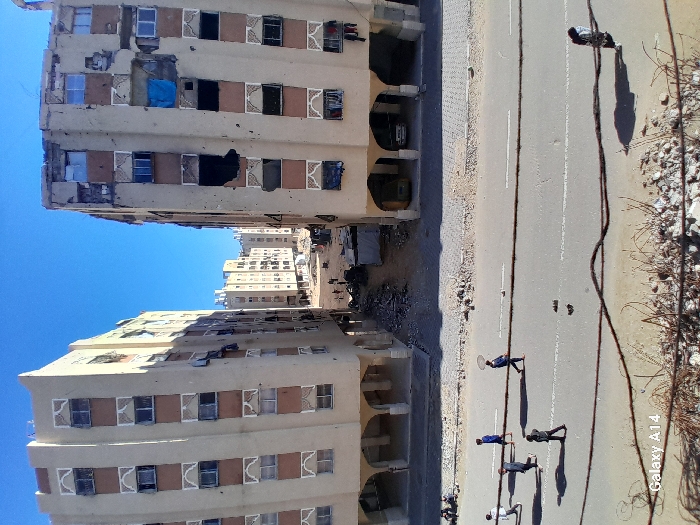 The image depicts a street scene in Khan Yunis, Palestinian Territory, flanked by two multi-story residential buildings under a clear blue sky. The photo is rotated 90 degrees counter-clockwise.
The two prominent buildings appear to be apartment blocks, characterized by light-colored facades with horizontal bands of brown and decorative patterns around many windows. Both structures show significant damage: sections of the facade are crumbling, revealing underlying construction materials and rebar, and many windows are broken or covered with dark fabric or blue tarps. Some upper-floor sections on the closer building on the left exhibit extensive damage. In the background, more similar buildings are visible, suggesting an urban residential area.
A paved road runs between the buildings, marked with a white center line and a dashed lane line. On the right side of the road, there is a dirt and rubble shoulder with scattered concrete pieces and what appear to be twisted metal wires or rebar. Several individuals are present on the road and shoulder. Five people are clearly visible in the mid-ground, casting long, distinct shadows across the road, indicating bright sunshine and possibly morning or late afternoon. They appear to be walking or standing.
Notable details include the widespread damage to the buildings, consistent with conflict or significant structural degradation. The bright blue sky and sharp shadows suggest a sunny day. In the bottom right corner of the original image, the text "Galaxy A14" is visible, indicating the device used to capture the photo. The overall scene portrays an environment marked by wear and potential conflict.