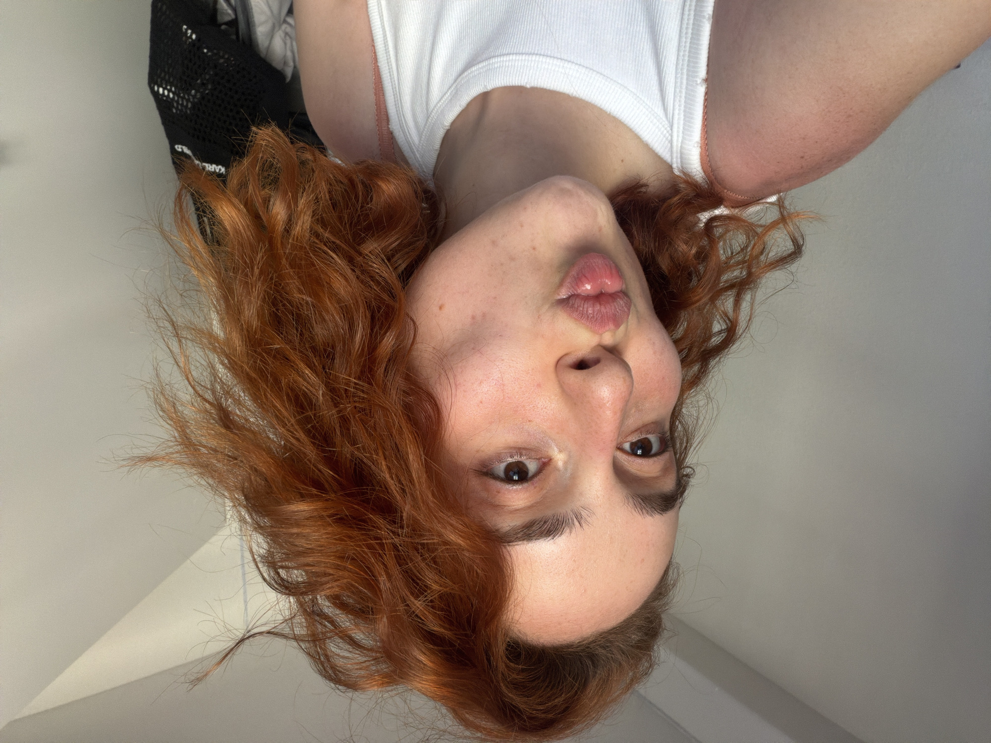The image is a close-up selfie of a young woman with auburn hair, puckering her lips. She is wearing a white tank top, and her right shoulder and upper arm show signs of sunburn. Her skin has freckles. The background is a plain, light-colored wall with some shadows, suggesting an indoor setting. There is no indication of the city of Strasbourg, France, in the image.
