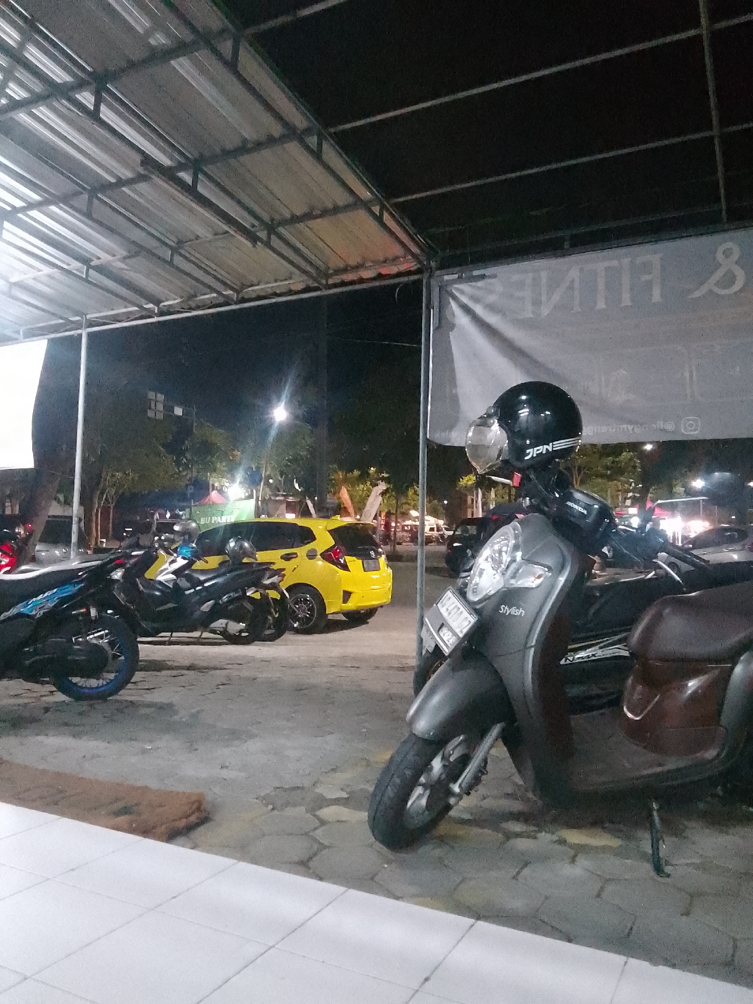 The image depicts a nighttime scene in what appears to be a parking area in Trenggalek, Indonesia. A gray scooter with a black helmet is prominently featured in the foreground, parked on a textured concrete surface. The scooter has a license plate that reads "B 6400 KZT". Behind it, several other motorcycles and a yellow car are visible. A white banner with text, partially obscured, hangs in the background, suggesting a commercial establishment. The overall environment is dimly lit, with overhead lighting and streetlights illuminating the area. Trees and buildings are visible in the distance. The scene suggests a public space, possibly a parking lot for a shop or event.