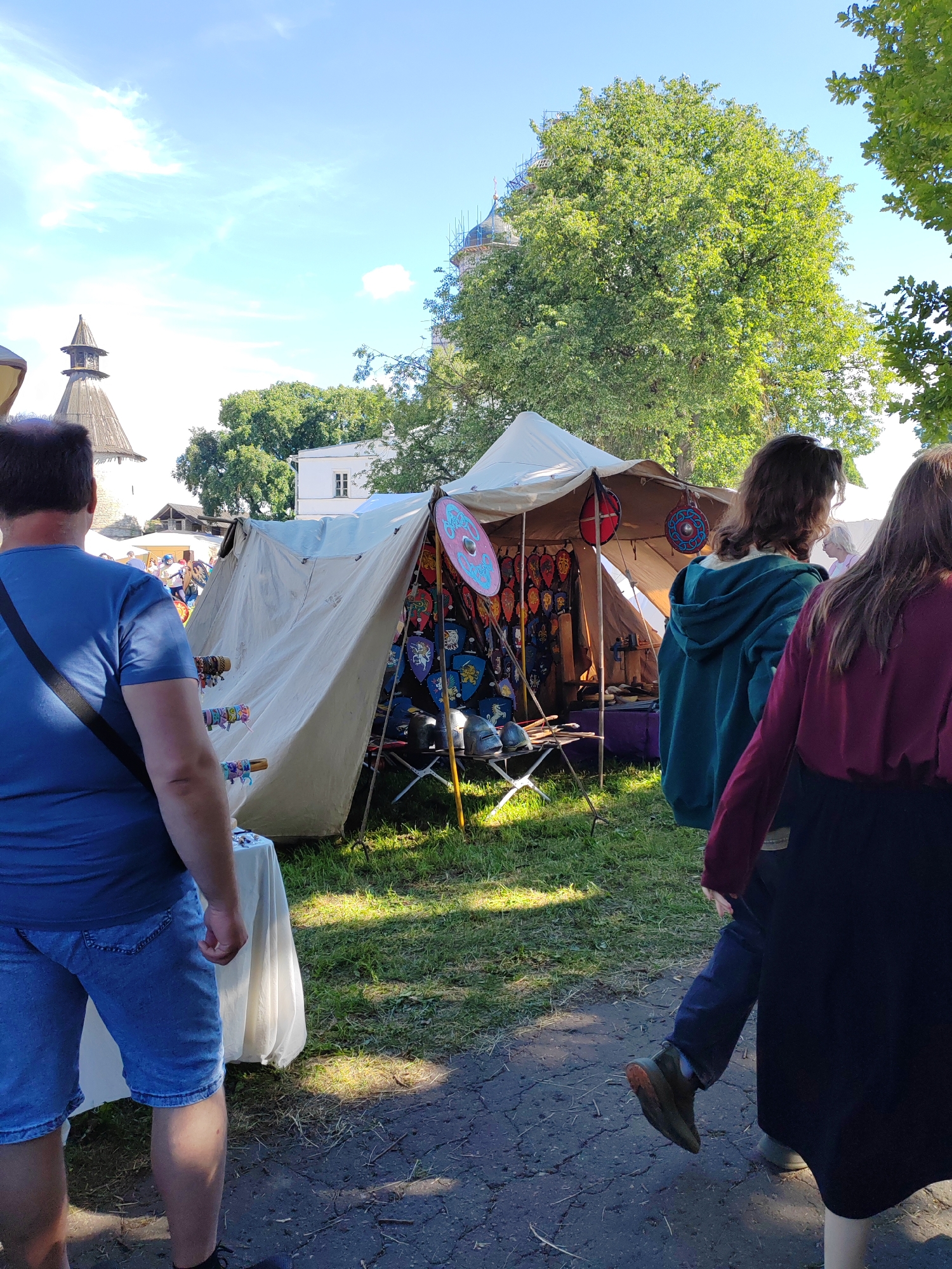 This media file captures a daytime outdoor scene at what appears to be a historical reenactment or festival. The primary subjects are several people, viewed from behind, walking through a grassy area. In the foreground, a man in a blue t-shirt and denim shorts is looking towards a large tent.
The tent, made of light-colored fabric, is adorned with numerous round shields, some bearing colorful emblems and patterns. Helmets and other historical items are also visible inside and around the tent, suggesting it is a vendor stall selling historical artifacts or costumes. Behind the tent, a large, mature tree with lush green leaves provides shade. In the background, the top of a stone structure with a pointed roof is visible, along with other buildings and trees, hinting at a historical site or a park. The sky is bright blue with a few scattered white clouds, indicating fair weather and likely a sunny day. The lighting suggests it is midday. The overall atmosphere seems relaxed and recreational, typical of a public event. No visible text is present in the image.