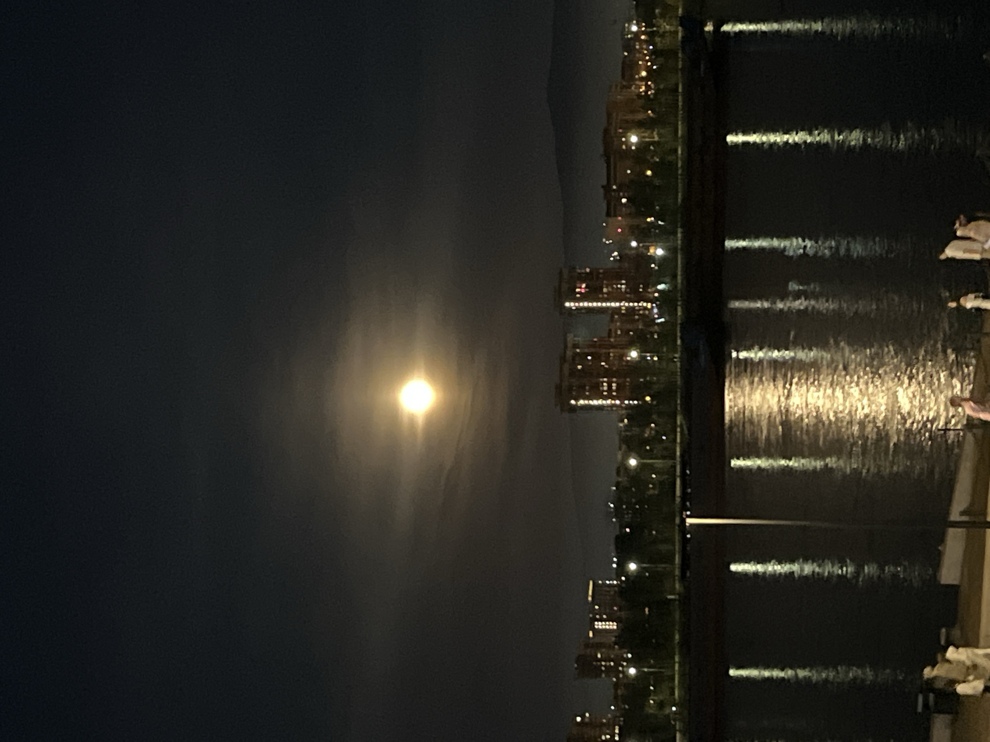 This media file presents a striking nighttime landscape, likely captured in Krasnoyarsk, Russia, as suggested by the context. The photograph is oriented vertically, showing a horizontal scene when correctly viewed.
The central and most prominent feature is a bright, full moon, positioned in the upper center of the dark night sky. It emits a powerful, luminous glow that creates a halo effect and casts a wide, shimmering reflection path across the calm body of water below. The sky itself is a deep, dark blue-black, with subtle hints of clouds or atmospheric haze around the moon. In the distance, faint, dark outlines of hills or mountains are visible against the skyline.
Beneath the moon and stretching across the midground is a city skyline. Numerous buildings, possibly residential or commercial, are illuminated with various lights, creating a warm, sparkling array of yellow and orange points of light. These city lights, along with the moon, are clearly reflected as elongated, wavy streaks on the dark, placid surface of the water, which appears to be a river or lake.
On the immediate right side of the image (when oriented correctly), a dark shoreline or embankment is visible. Along this edge, there are some indistinct, light-colored shapes or structures, possibly indicating part of a dock, steps, or other riverside features. No specific activity or visible text can be identified in the image. The overall impression is one of serene urban beauty under a brilliant moonlit sky.