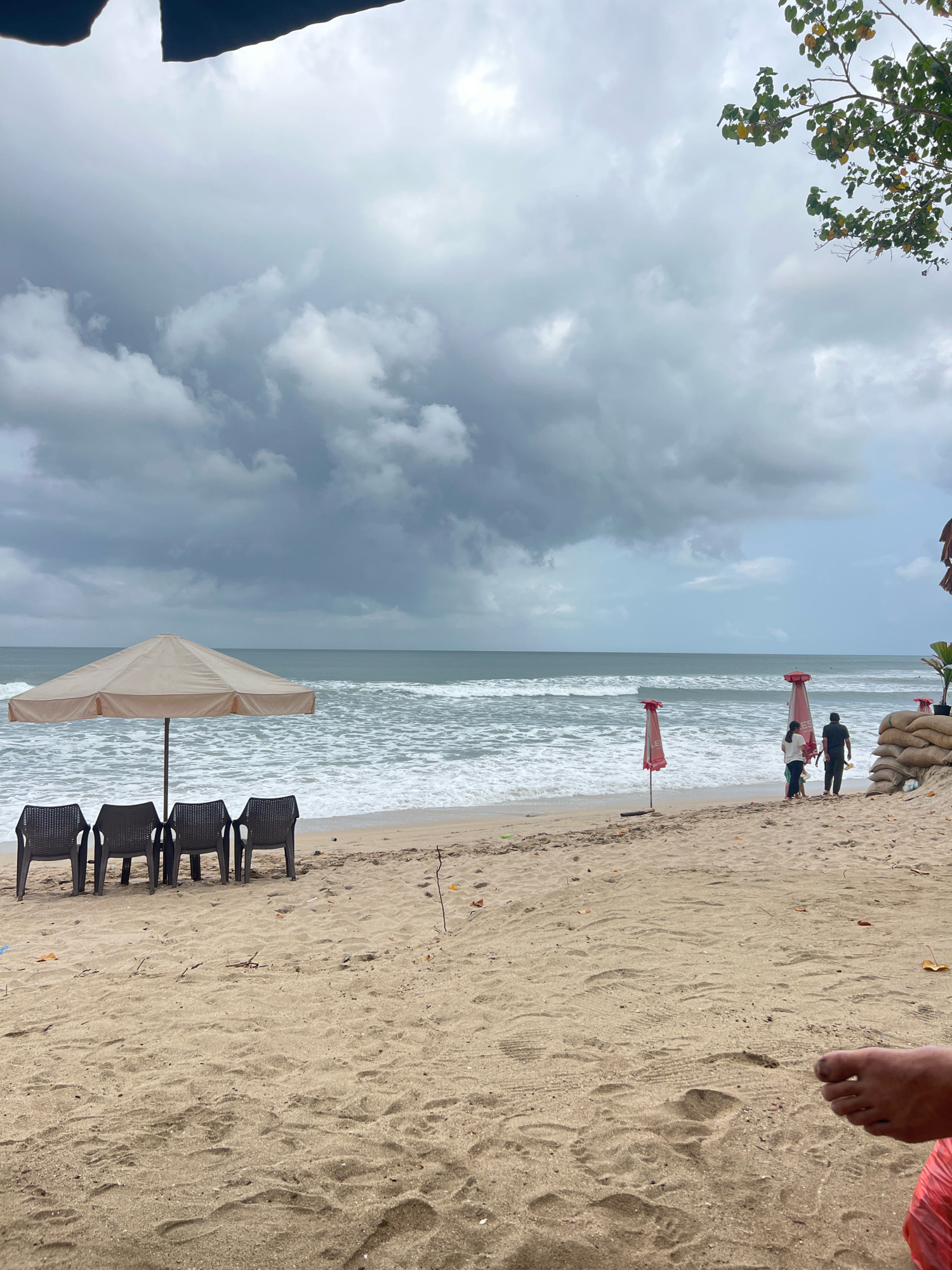 This image depicts a cloudy day on Kuta Beach, Bali, Indonesia. In the foreground, there are four empty dark plastic chairs arranged in a row beneath a large, light-colored beach umbrella. The sand is visible throughout the foreground.
Further back on the beach, a couple with a child are walking towards the ocean. Two red beach umbrellas are partially visible behind them. To the right, a stack of sandbags is present, with some vegetation growing around it. The ocean is in the background, with visible waves and a foamy surf. The sky is overcast with heavy, gray clouds, and a tree branch with green leaves is visible in the upper right corner. The overall scene suggests a beach resort atmosphere, likely during the off-season or a period of inclement weather.