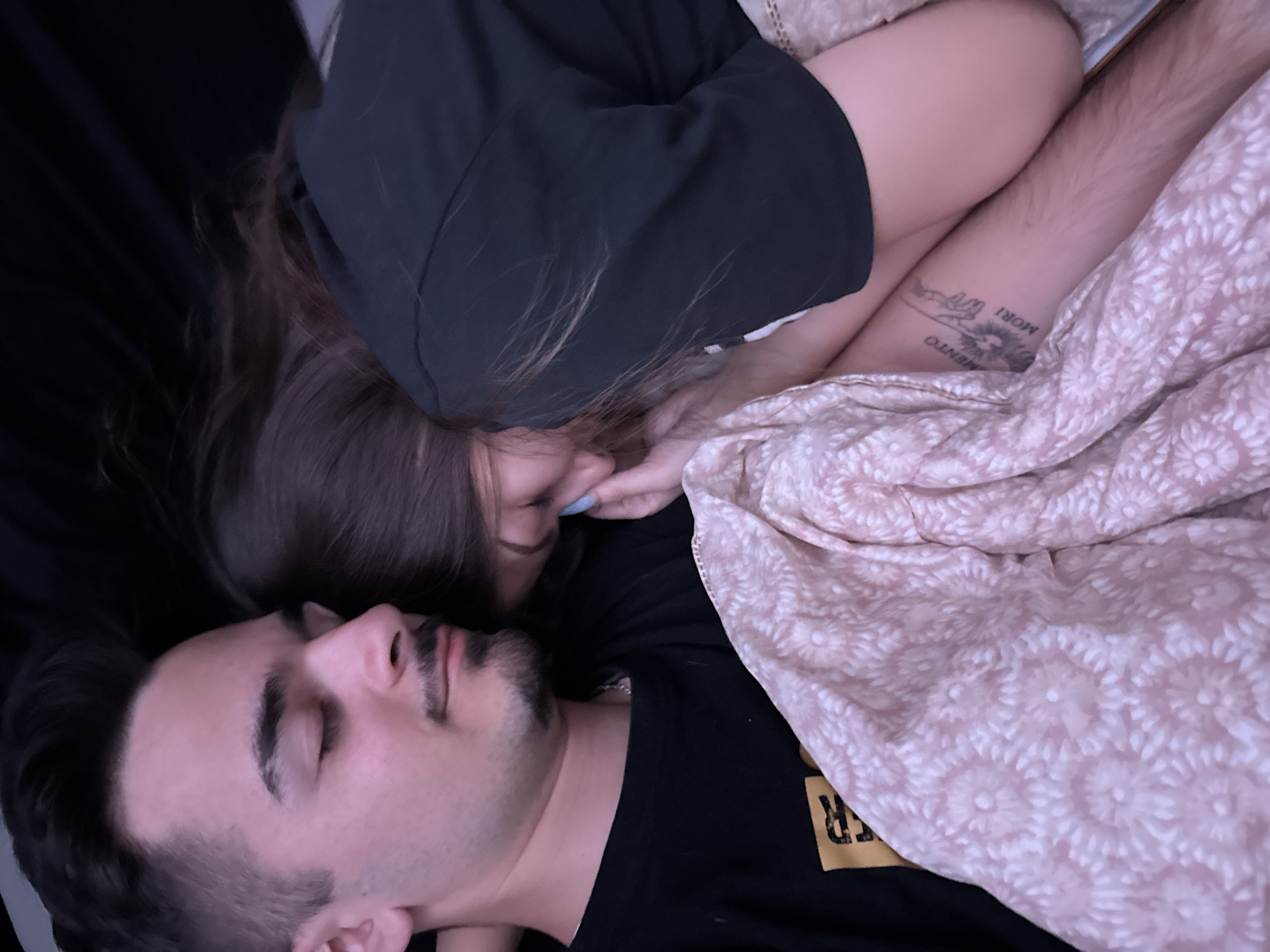 Two individuals are depicted in a close embrace, resting or sleeping in a dimly lit room located in Sofia, Bulgaria. The individual on the left, a male-presenting person with dark hair and a short beard, is seen in profile with eyes closed, wearing a dark T-shirt with a visible yellow/orange graphic and a silver chain. The individual on the right, a female-presenting person with long dark hair, is nestled against the first individual's chest, also wearing a dark T-shirt. Her face is partially obscured by her hair and position. Her fingernails are painted a light blue color. A light pink blanket with a repeating white floral pattern covers both individuals from the lower torso downwards. The female-presenting individual's right upper thigh is visible, displaying a tattoo that includes what appears to be two hands and text, with the partial inscription "MENTO MORI." The background is uniformly dark, suggesting a bedroom environment under low light conditions.