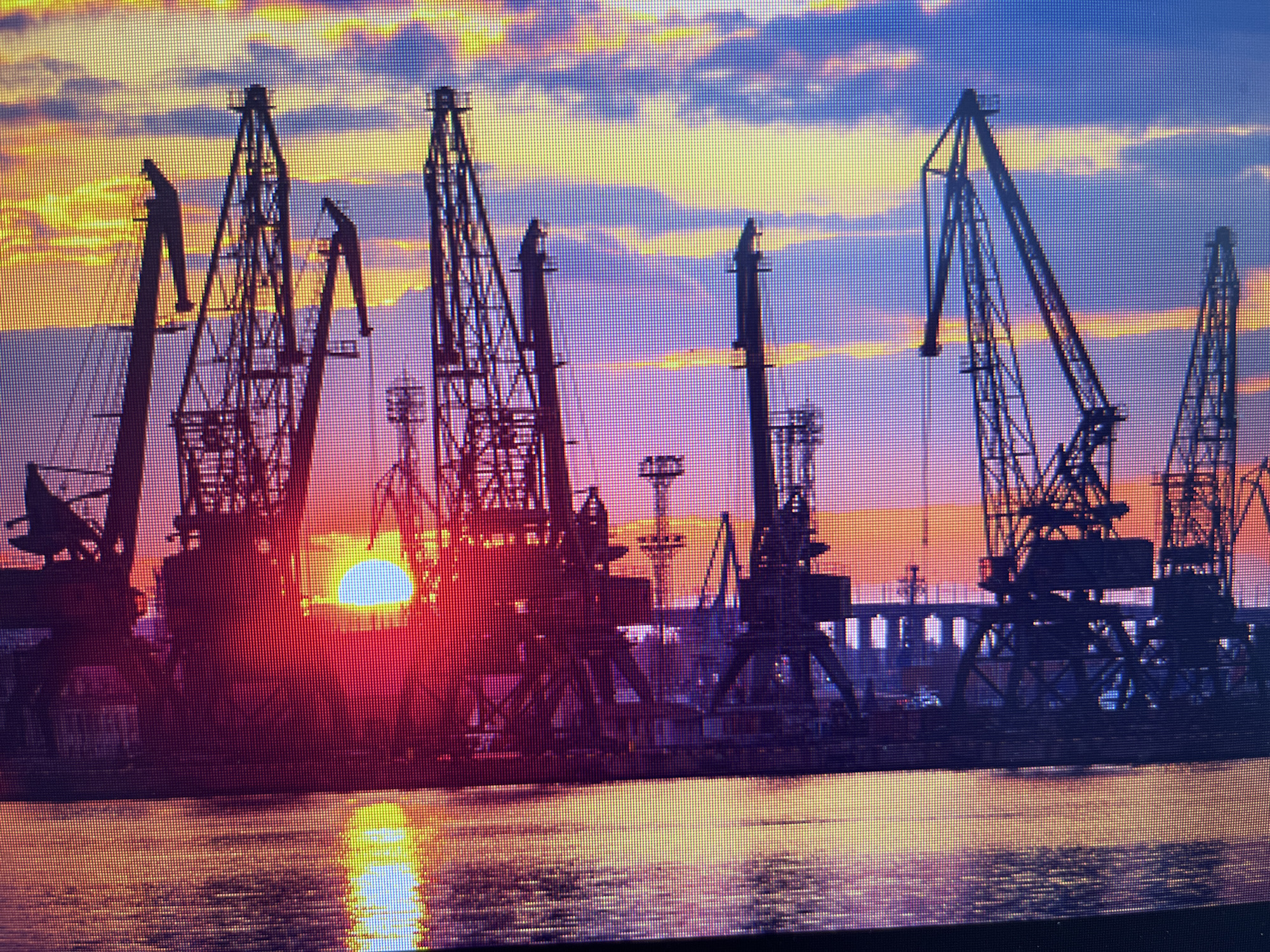 The image depicts a port scene in Varna, Bulgaria, at sunrise or sunset. Several large harbor cranes dominate the foreground, silhouetted against the sky. The sun is partially obscured by the cranes, creating a bright glare. The water in the foreground reflects the colors of the sky. In the background, structures and lights suggest the presence of a bridge or other buildings. The sky transitions from blue to yellow and red hues, indicating the time of day.