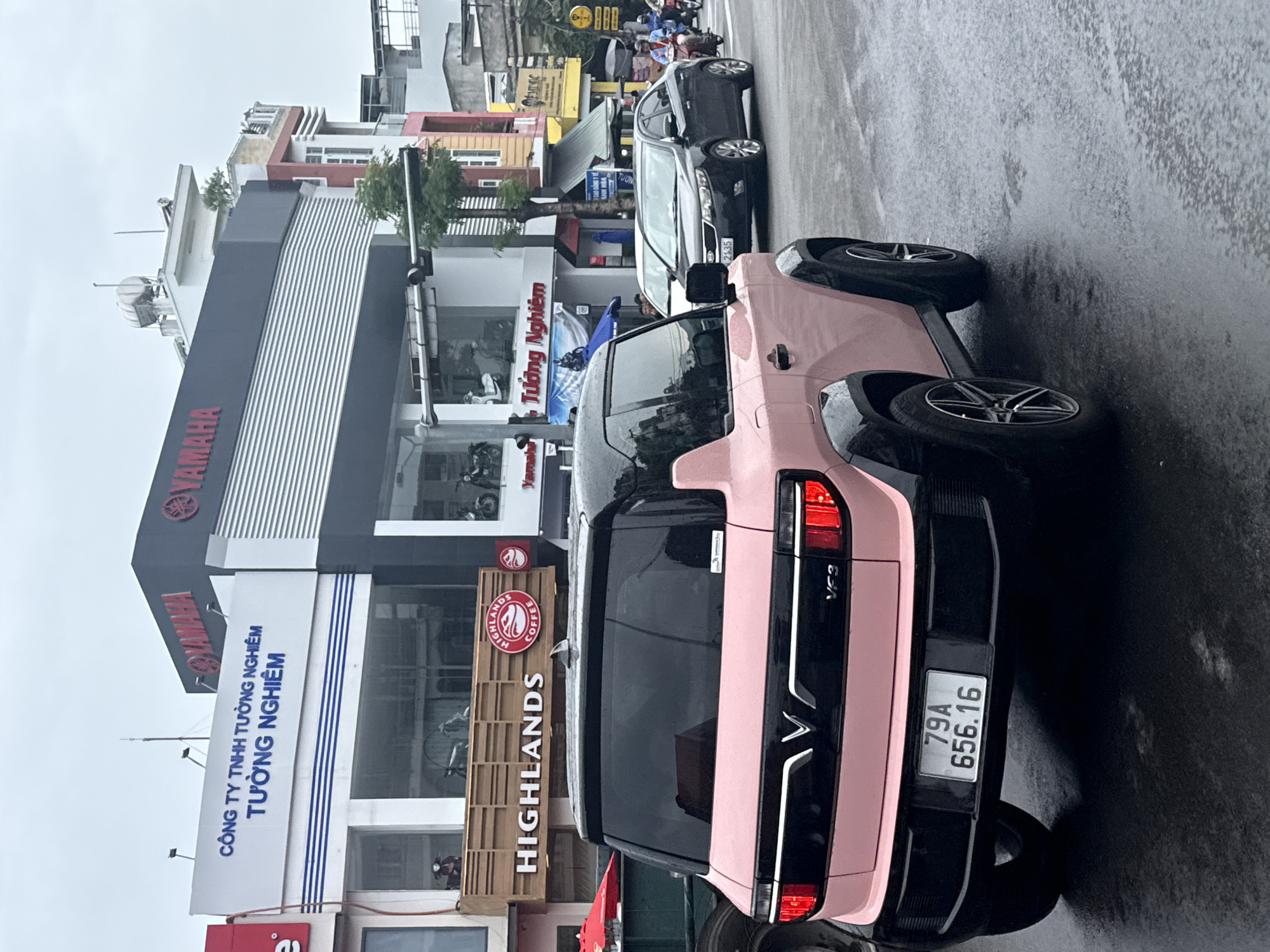 In Nha Trang, Vietnam, on a wet asphalt street, a pink VinFast VF3 car is parked in the foreground. The car's rear is visible, featuring a black bumper and tailgate with a "VF3" emblem. A license plate reading "79A 656.16" is affixed to the rear. To the right of the VF3, a black sedan is seen driving. In the background, a building with multiple levels displays signs for "YAMAHA" and "HIGHLANDS COFFEE," indicating commercial establishments. The sky is overcast, suggesting a cloudy day.