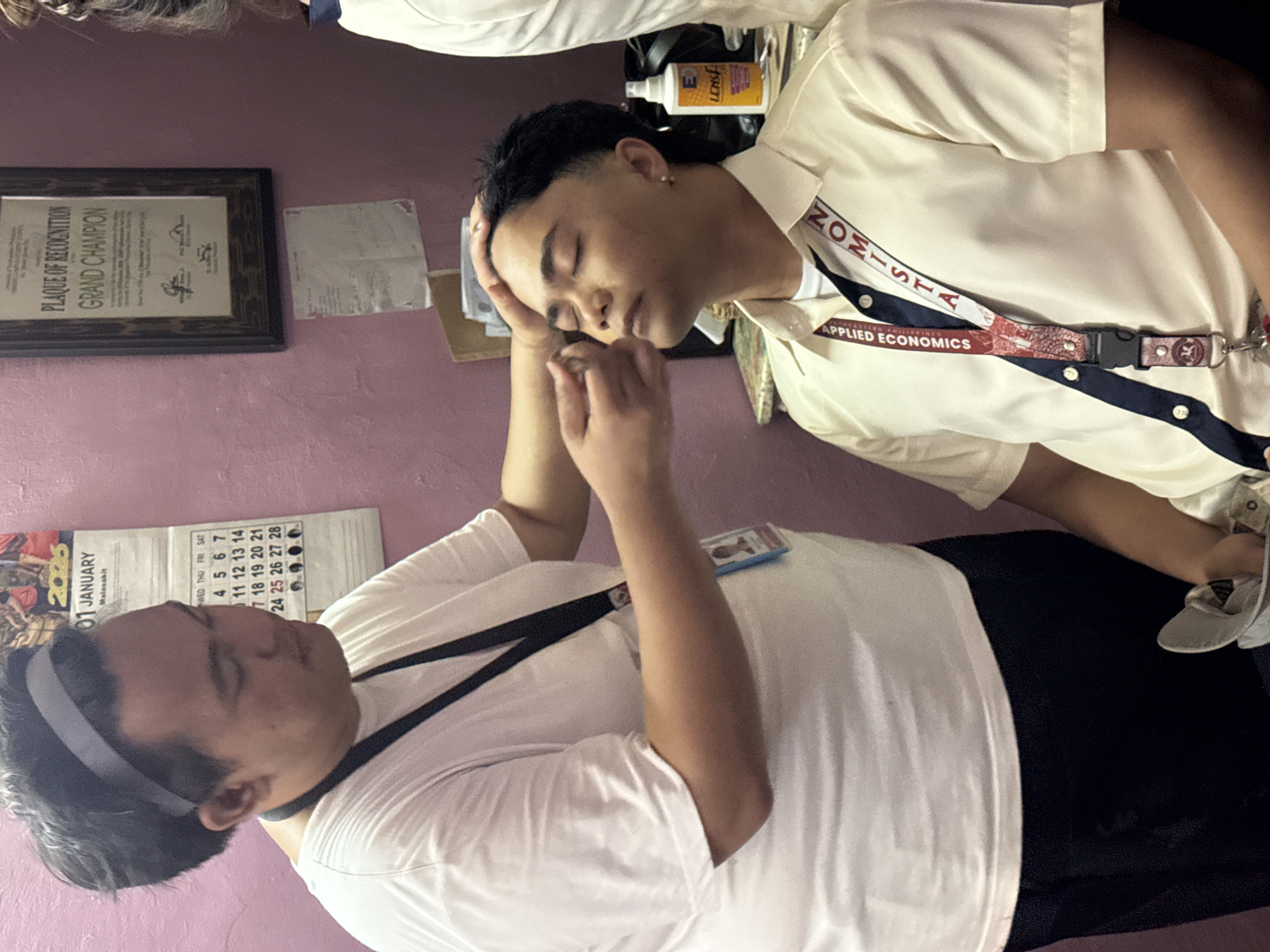 In an indoor setting with pink walls, a person wearing a white t-shirt and a grey headband is applying makeup to another person who is wearing a light yellow collared shirt with a navy blue accent and a lanyard that reads "APPLIED ECONOMICS". The person receiving makeup has their eyes closed and is wearing an earring. A calendar showing January 2020 is visible on the wall behind the makeup artist. To the right, a plaque that reads "PLAUQE OF RECOGNITION GRAND CHAMPION" is also mounted on the wall. A bottle labeled "LENS" is on a surface to the right of the person receiving makeup. The image does not provide specific details about the location in Davao City, Philippines.