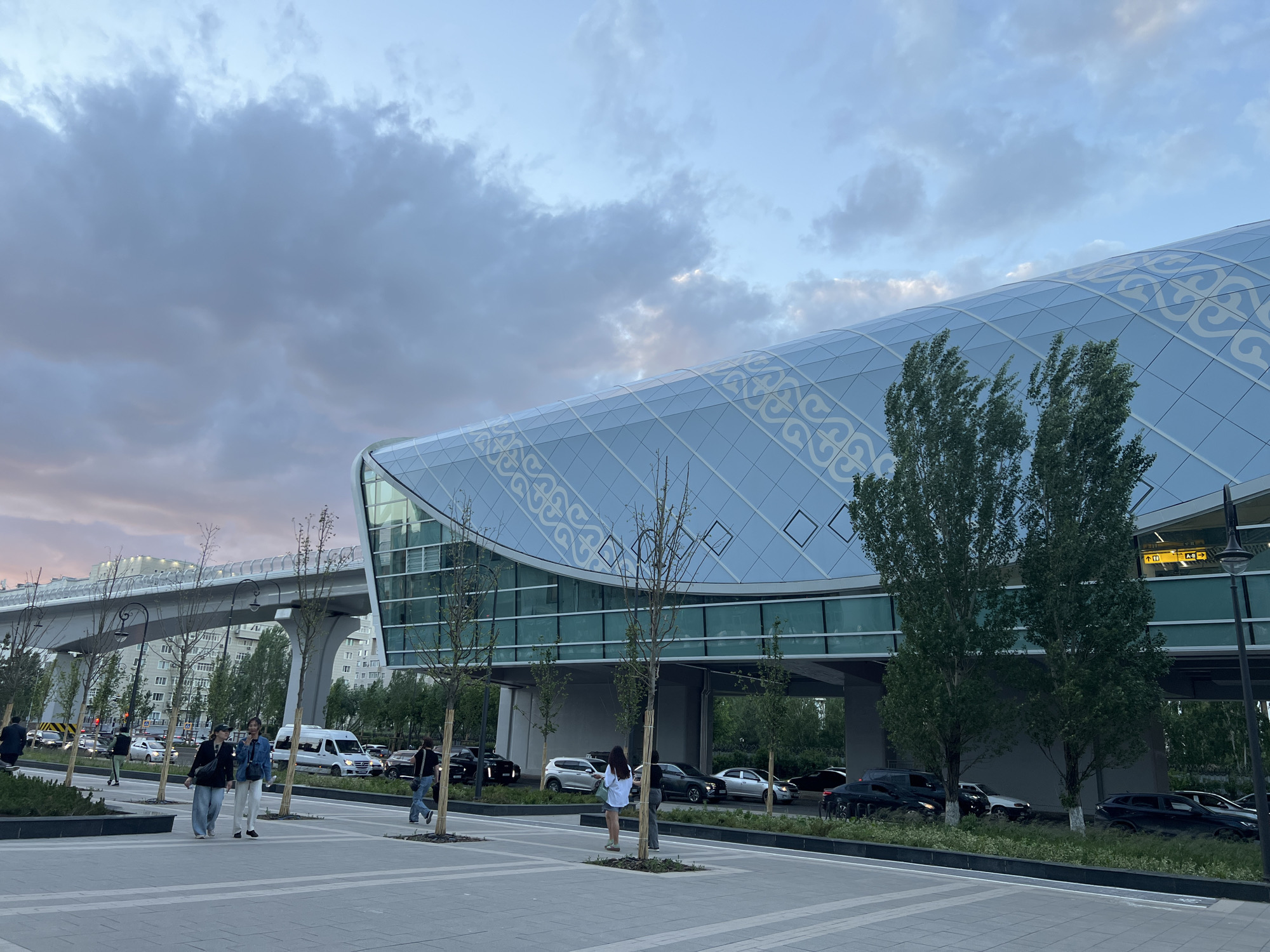 The image depicts a modern building with a curved glass facade, adorned with a pattern reminiscent of traditional Kazakh designs, in Astana, Kazakhstan. A pedestrian plaza is in the foreground, featuring trees and people walking. A raised roadway, likely part of the city's infrastructure, is visible in the background. Cars are parked near the building. The sky is overcast with hues of grey and pink, suggesting either sunrise or sunset. People are casually dressed, engaging in everyday activities.