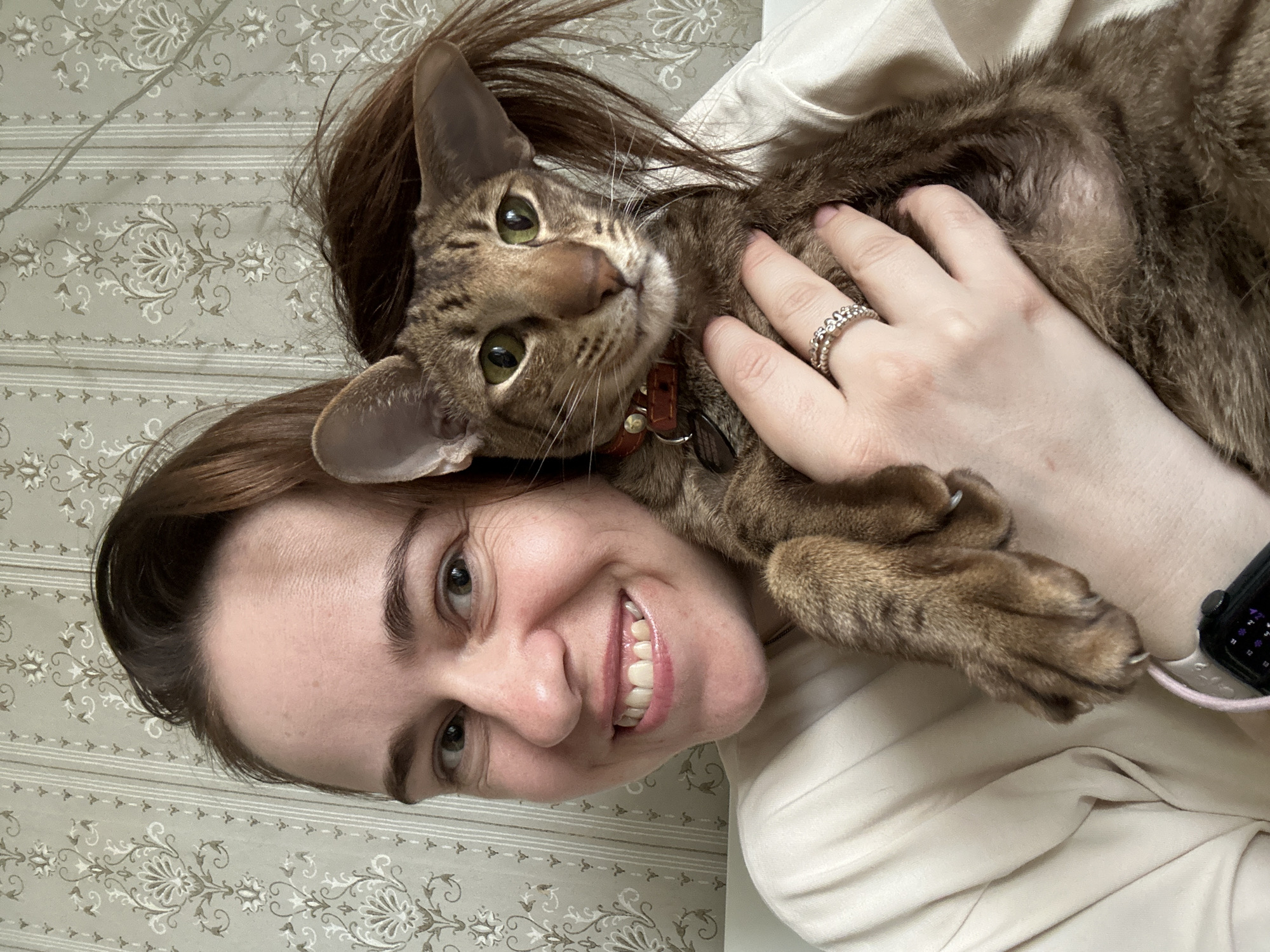 An indoor shot captures a close-up of a woman and a cat, both looking directly at the camera with a slight upward gaze. The image is oriented vertically, with the woman's head and the cat's upper body visible as if they are lying down together.
The woman, with fair skin, brown hair, and green eyes, displays a wide, cheerful smile showing her teeth. She wears a light-colored top, possibly cream or white. On her left ring finger, she sports a silver-colored ring featuring a delicate floral or interconnected loop design. A smartwatch with a light-colored band (possibly pink or light grey) is visible on her left wrist, displaying small, indistinct white icons and numbers on a dark screen. Her right hand is gently placed on the cat, indicating affection.
The cat, likely a Bengal or similar breed, has striking brown spotted/striped fur (tabby pattern) and bright green eyes. It is wearing a thin red collar with a small, unreadable tag. The cat appears calm and attentive, nestled against the woman's chest or shoulder.
The background features a light greyish-green wallpaper or fabric with an elegant pattern of white and beige floral motifs and subtle horizontal stripes. The lighting is bright and even, suggesting a well-lit indoor environment, likely during the day. The overall mood is warm and affectionate. This scene takes place in Moscow, Russia.
