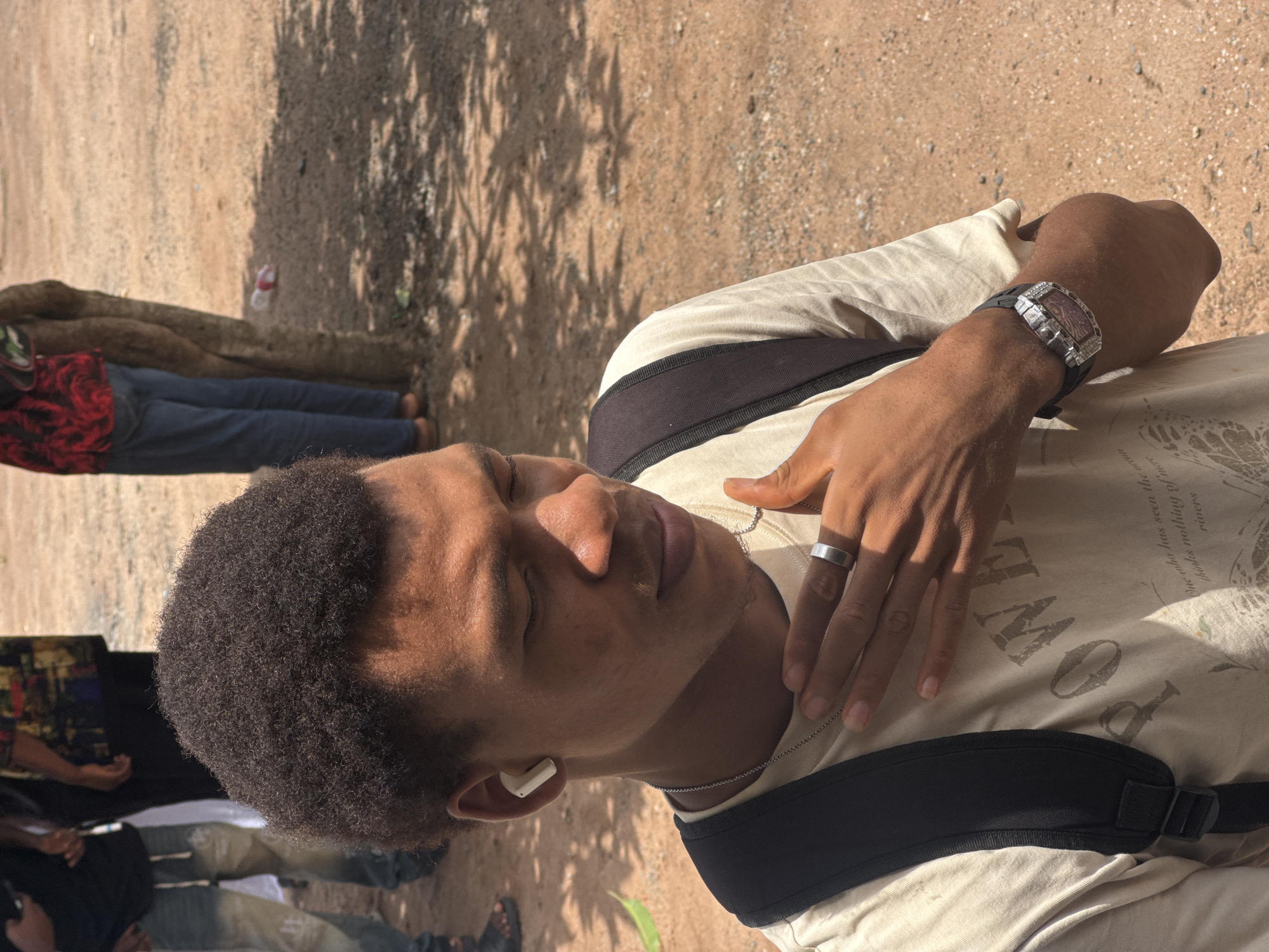 An outdoor, eye-level, medium-shot photograph captures a young Black man from the chest up. He is looking down and to his right, with his eyes closed and his head tilted slightly. A white earbud is visible in his left ear. His right hand is raised to his neck, with his thumb resting on his collarbone and his fingers extended downwards. He wears a silver ring on his right index finger. He is also wearing a light-colored t-shirt with a dark-colored backpack strap visible over his left shoulder. The t-shirt has an emblem or text printed on it. He is wearing a watch on his left wrist. The background is blurred and shows a dirt path with scattered pebbles, some shadows from trees, and a few people in the distance. One person in the background is wearing a red and black patterned top and blue jeans. Another person to the left is wearing blue jeans and a black t-shirt. The scene is set in Ihiagwa, Nigeria.