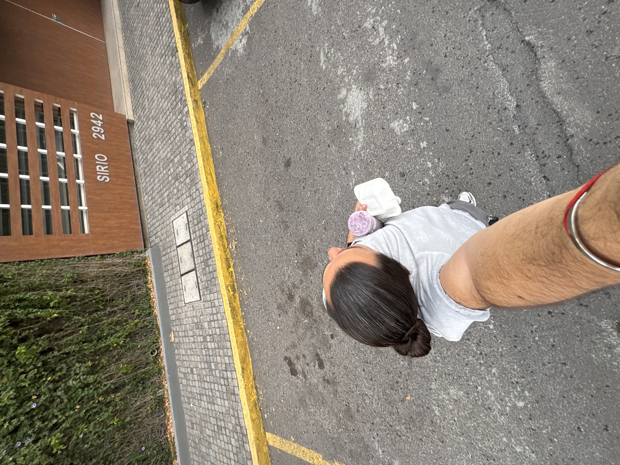 A man with dark hair pulled back into a bun takes a selfie at street level. He is holding a takeout container and a purple beverage. He is standing on a gray asphalt surface marked with yellow lines. Adjacent to the pavement is a walkway with gray pavers. Next to the walkway is a building facade with the address "Sirio 2942" visible. In front of the building, dense green foliage is planted along the sidewalk. The location is the city of Puebla, Mexico.