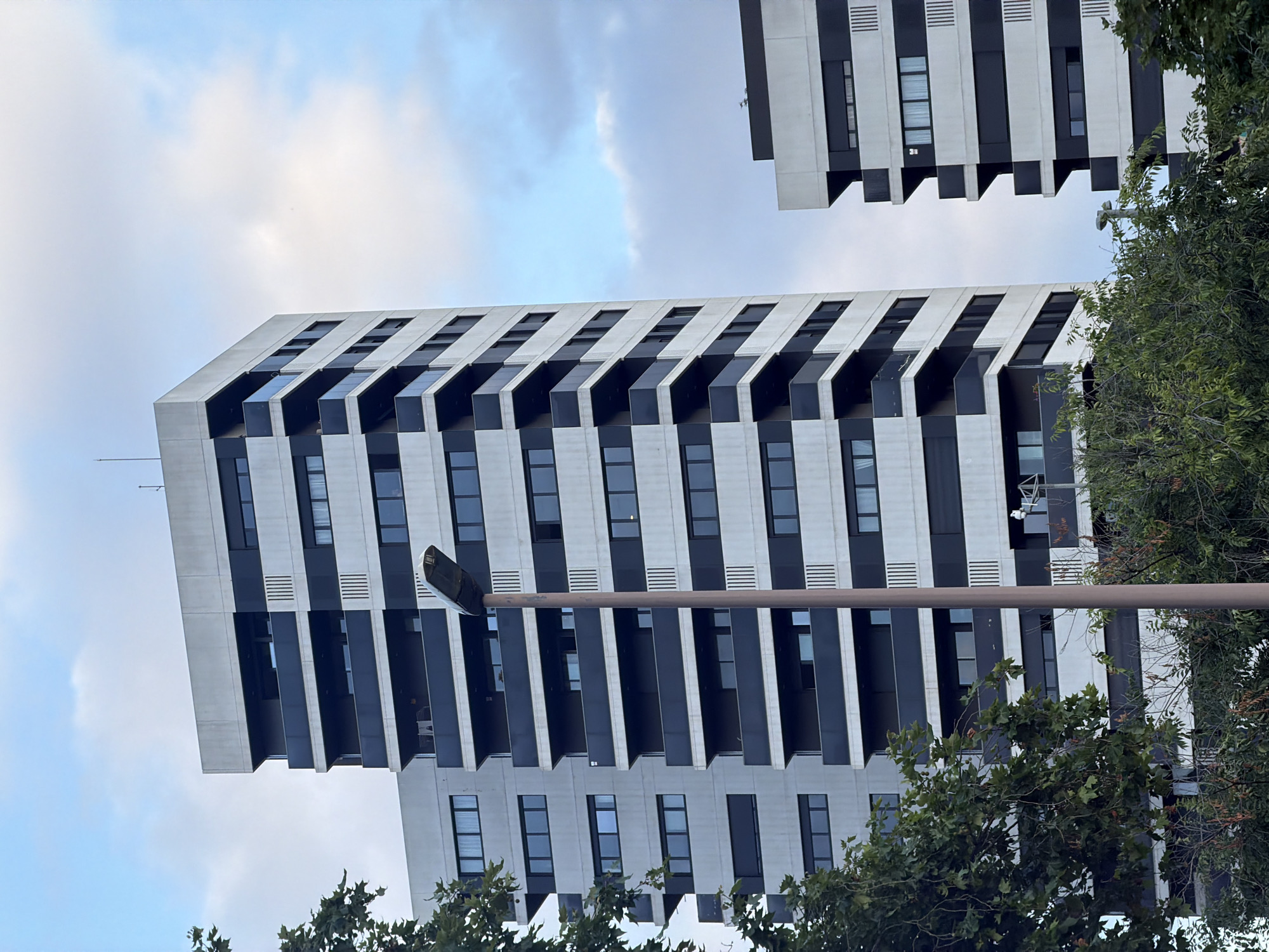 This media file features two modern apartment buildings with striking architectural designs. The buildings are characterized by a repetitive pattern of windows and balconies, with alternating bands of white and dark grey or black. The balconies are recessed, creating a sense of depth and shadow. The building in the foreground is angled and appears to be leaning or viewed from a low angle, emphasizing its verticality. The second building, seen in the upper right corner, is positioned at an angle relative to the first, suggesting a complex urban landscape.
The scene is set against a cloudy sky, with diffused light suggesting it is daytime, possibly late afternoon or early morning given the cloud cover. The presence of lush green trees in the foreground and partially obscuring the lower portions of the buildings hints at a vibrant urban environment, possibly a park or well-maintained residential area. The overall mood is serene and modern, highlighting the contemporary architecture and natural elements.
There are no people visible in the image. The main objects are the two buildings and the trees. A street lamp is prominently visible in the foreground, running horizontally across the frame and partially obscuring the view of the buildings.
While no explicit text is legible on the buildings themselves, the architectural style and the presence of trees suggest an urban setting, possibly residential, in a city like Barcelona, Spain, as indicated by the location context.