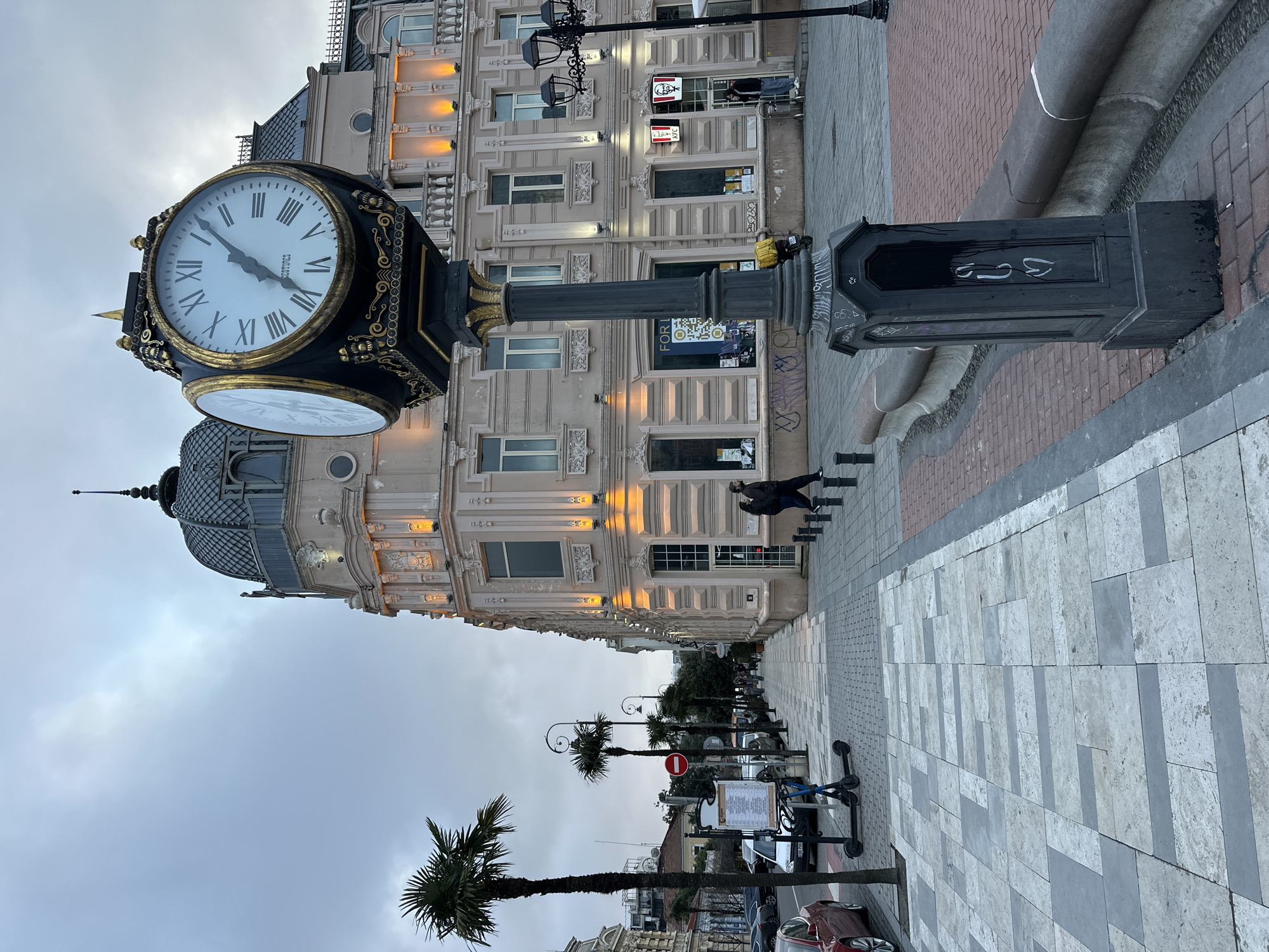 The image shows a street in Batumi, Georgia. A large, ornate, black and gold clock with two faces stands on the sidewalk. A building with decorative elements and warm lighting is behind the clock. The street has palm trees and streetlights, and some people are visible. The sky is overcast. The sidewalks are paved with light grey stone, and there are a few parked cars. A KFC restaurant sign is visible on the building behind the clock. A pedestrian is walking on the sidewalk.