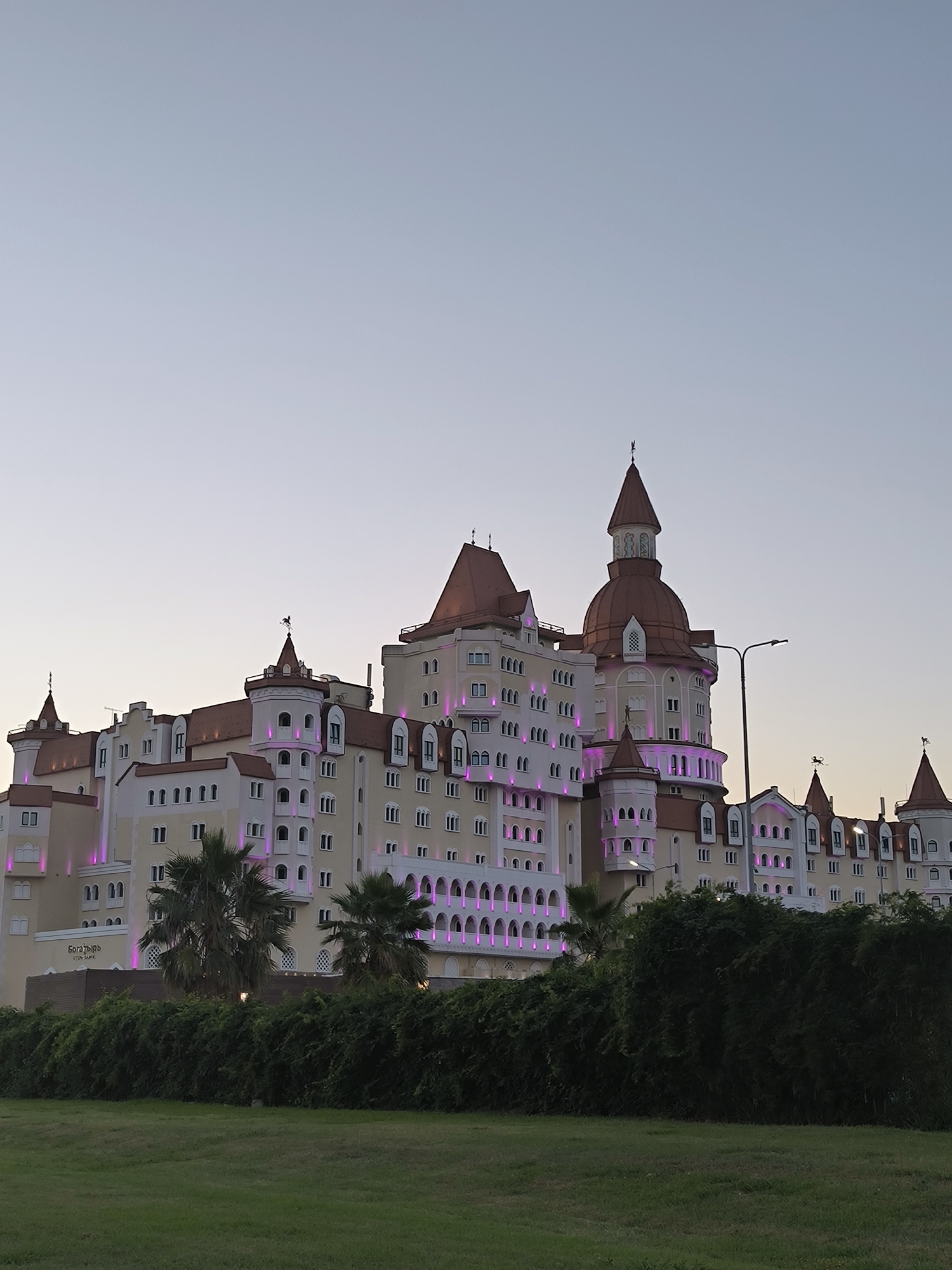 The image depicts the Radisson Collection Paradise Resort & Spa, Sochi, a large, ornate hotel complex with a castle-like architectural style, located on the Black Sea coast in the Adler district of Sochi, Russia. The hotel features multiple towers with red-tiled roofs, arched windows, and decorative elements. Pink lights illuminate the facade of the building. In the foreground, there is a lush green lawn and a dense hedge of dark green foliage. Two palm trees are visible on the left side of the hotel. The sky is a pale, clear gradient of blue and grey, suggesting either early morning or late evening. There are no people or animals visible in the scene.