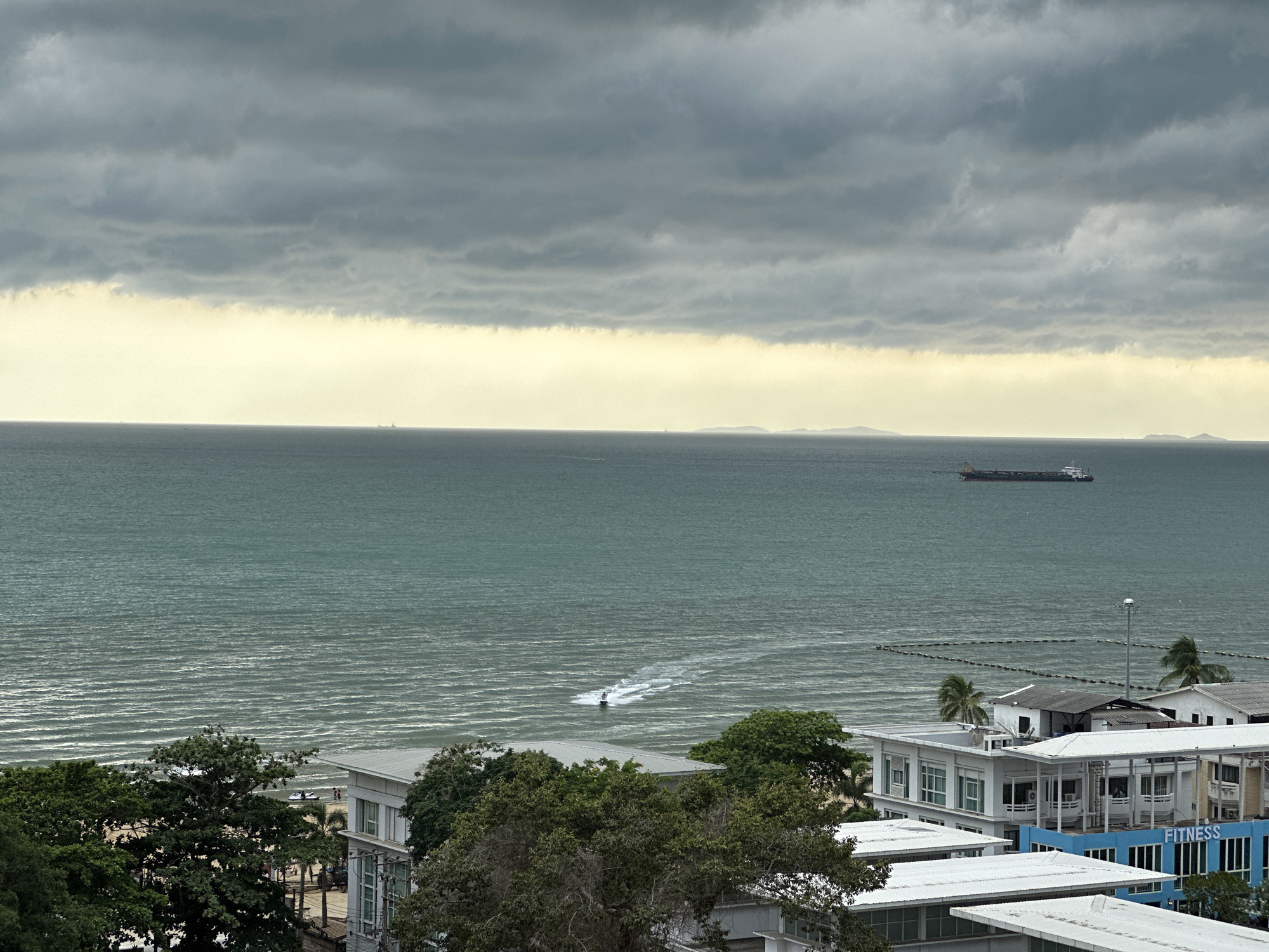 The image shows an overcast day over the ocean in Pattaya City, Thailand. A cargo ship is visible on the horizon, to the right of the frame, and a small island is visible in the far distance, toward the middle of the frame. A personal watercraft leaves a trail in the water near the shore, at the center of the frame. Buildings with white and blue facades are visible in the foreground, along with green trees.