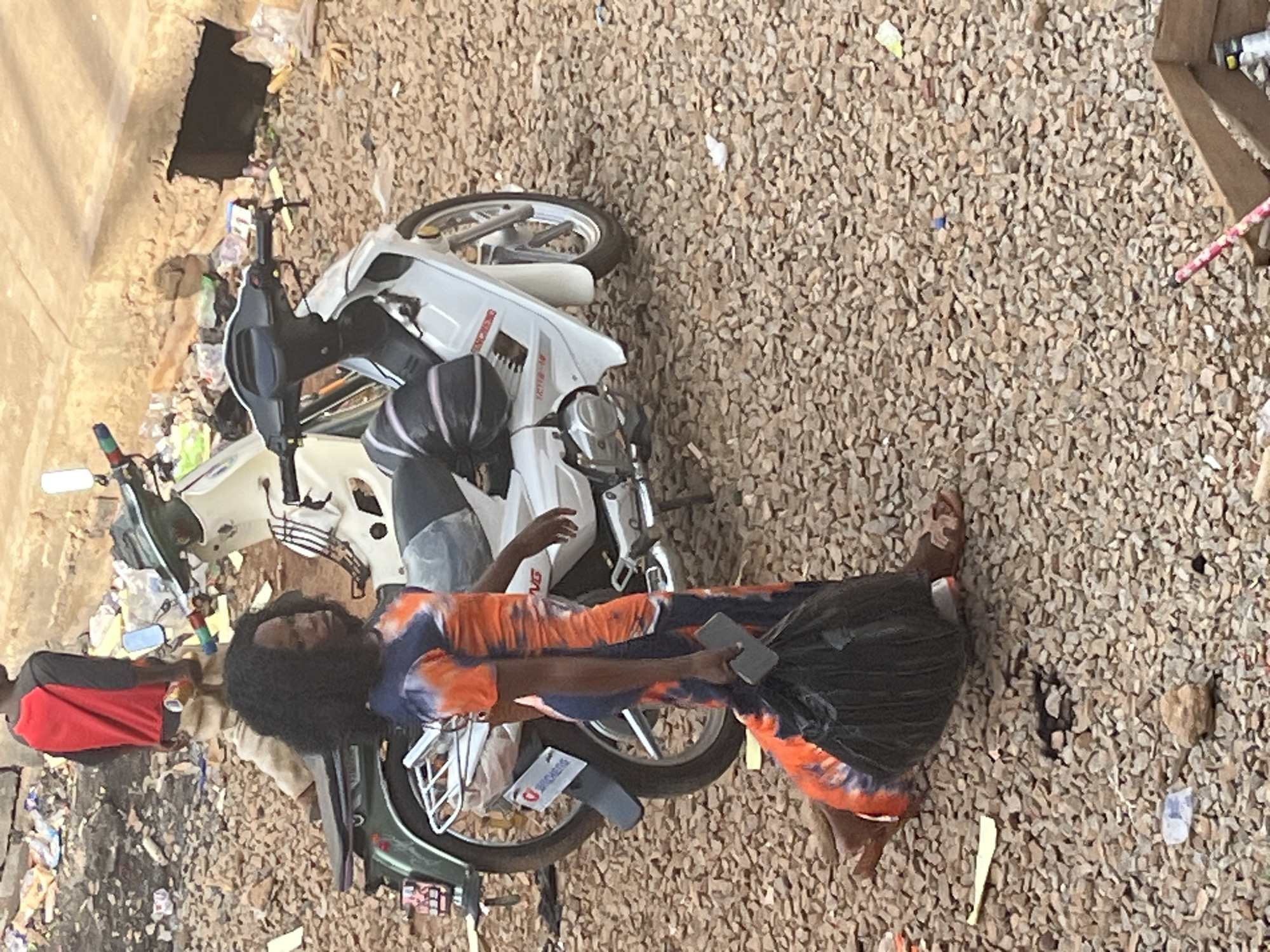 In an outdoor setting on a gravelly surface in Jos, Nigeria, a Black woman with dark, curly hair is depicted walking. She is wearing a tie-dye dress with orange and dark blue patterns and appears to be carrying a large black plastic bag in her left hand and a grey mobile phone in her right hand. She is wearing brown sandals. Behind her, a white motorcycle is parked, and to the left, another motorcycle with a green seat is partially visible. In the background, a young person wearing a red and black long-sleeved shirt and beige pants is standing, holding a cylindrical object. The ground is covered with small stones and scattered debris, including plastic bags and wrappers, suggesting a somewhat unkempt environment.
