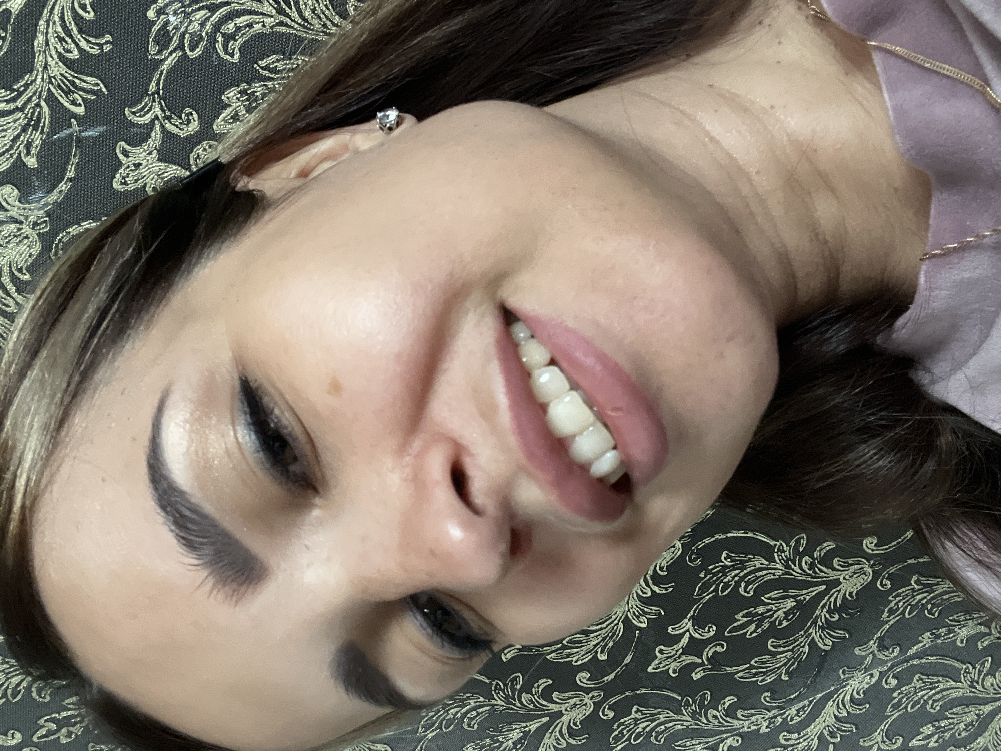 A close-up, low-angle indoor portrait shows a woman with long, dark brown hair, her face turned to the right, smiling and showing her teeth. She wears a light pink top and a gold chain necklace. She has on eye makeup and a silver stud earring in her right ear. The background is a dark, patterned wallpaper. The photo appears to have been taken by the subject herself.