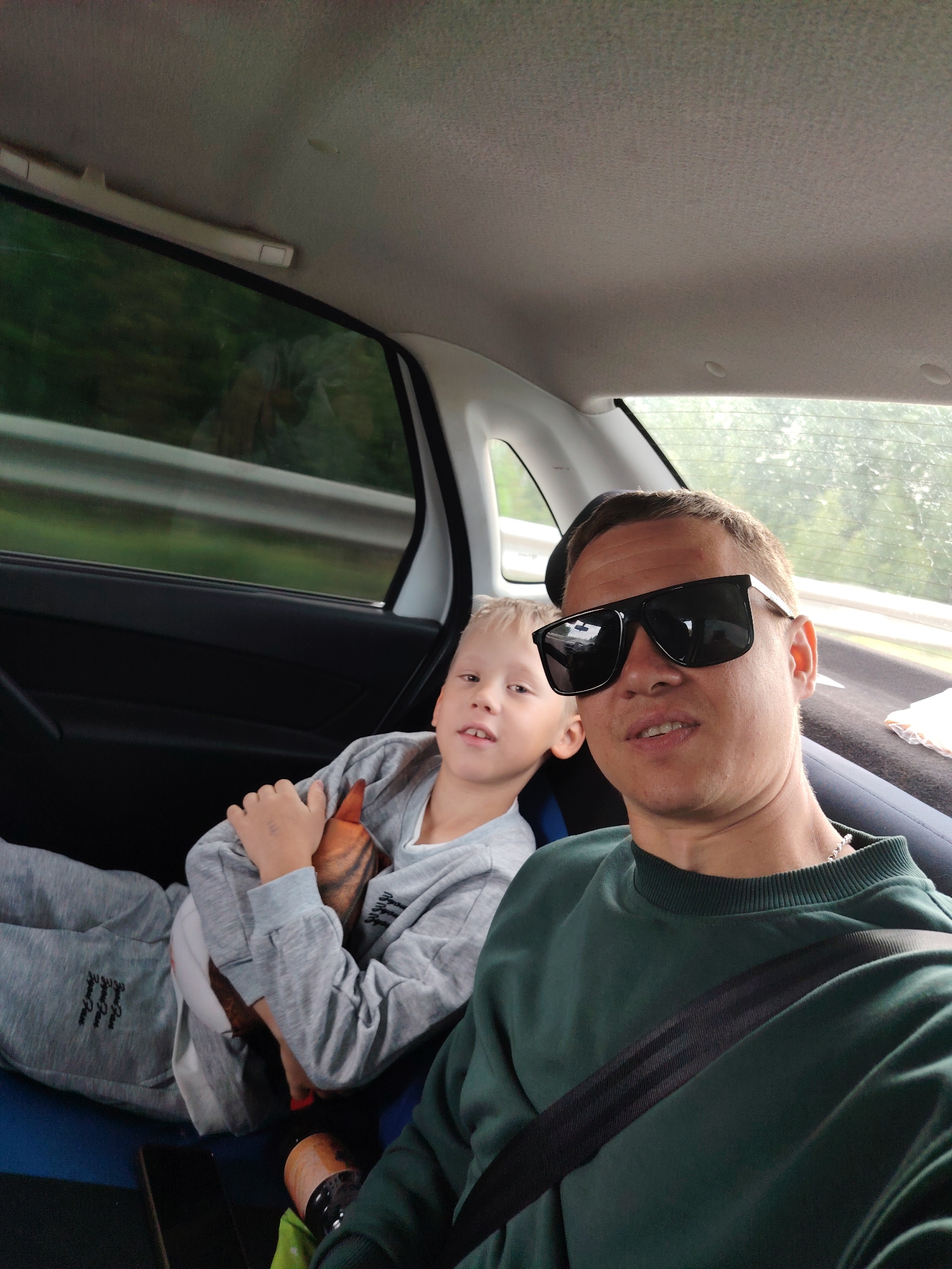 The image shows a selfie taken from inside a car. A man wearing dark sunglasses and a green sweater with a black seatbelt across his chest is in the foreground. Behind him, a boy with blond hair is sitting in the backseat, wearing a gray hoodie and pants. The boy is holding a stuffed animal. The car is moving, as suggested by the blurred scenery outside the window. Based on the geolocation Неволино, Russia, the photo was likely taken within the vicinity of that location.