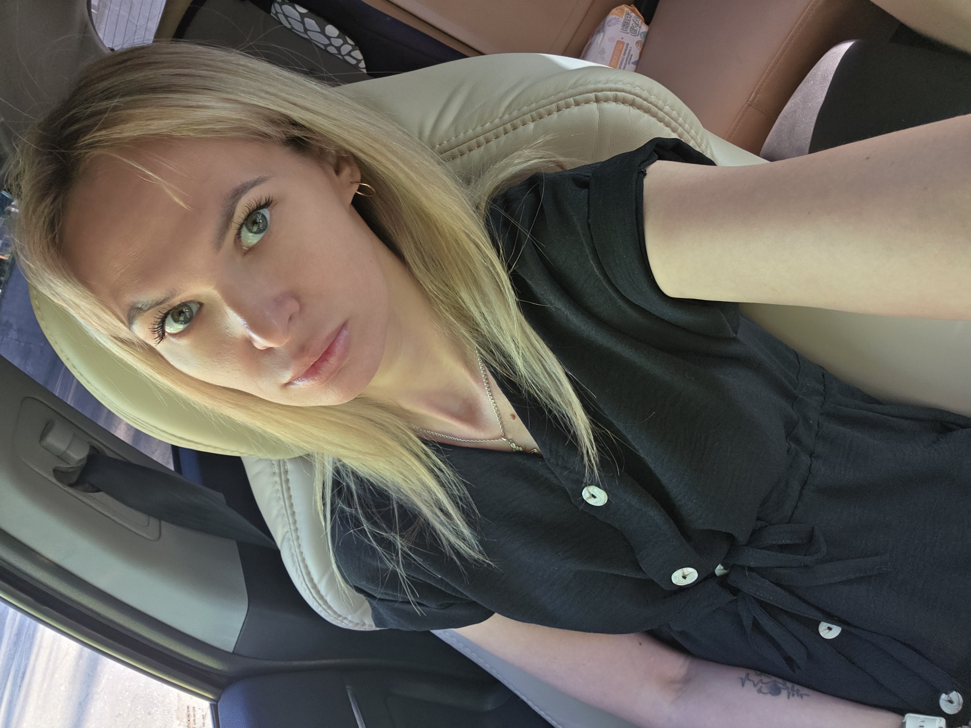 A blonde woman is sitting in the passenger seat of a car. She wears a black dress with white buttons and a silver necklace. She has green eyes and is looking at the camera. The car seat is beige leather. The car's interior is visible, including parts of the door and other seats. A package is visible in the background. The exact location in Chaikovsky, Russia is not discernible from the image.