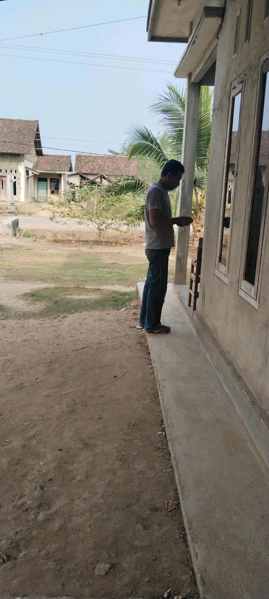 A man in a grey t-shirt and blue jeans stands on a concrete pathway in front of a building. He is looking down at something he is holding in his hands, possibly a phone. The building appears to be unfinished or under construction, with exposed concrete and wooden framing. In the background, several houses with tiled roofs are visible, suggesting a residential area. The ground is a mix of dirt and grass. Palm fronds from a nearby tree hang over the building. The sky is clear and pale blue. The scene depicts a casual moment in what appears to be a rural or semi-rural setting in Tanjung Ratu Ilir, Indonesia.