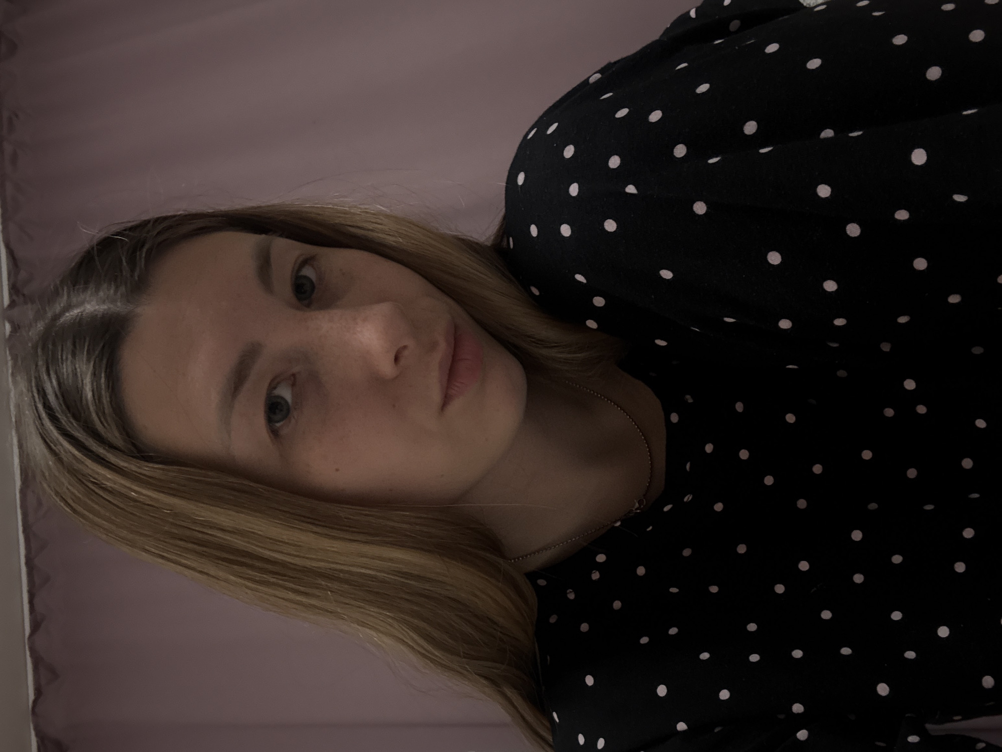 A fair-skinned woman with blonde hair is visible in a selfie-style photograph. She is wearing a black blouse with white polka dots and a delicate silver necklace. Her gaze is directed at the camera. The background features a light pink wall, and part of what seems to be a window frame is visible on the left. The image does not provide any information about the location in Moscow, Russia.