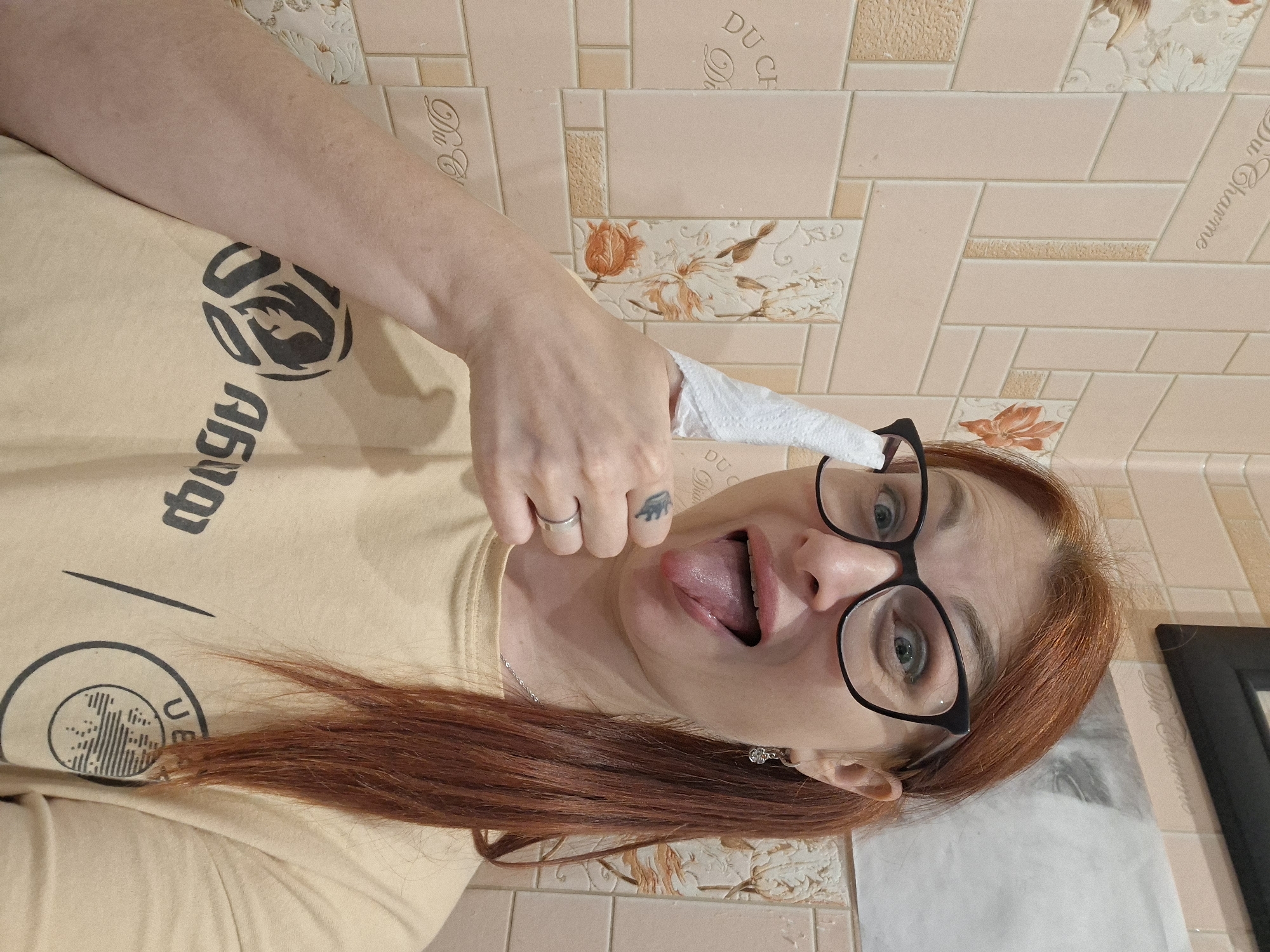 A woman with red hair, wearing glasses and a beige t-shirt, sticks out her tongue while holding a tissue to her thumb. She is standing in front of a tiled wall with floral patterns. The wall has tiles arranged in a brick pattern, with some tiles featuring intricate floral designs and others having decorative text. The lighting in the scene is consistent with indoor ambient lighting, possibly from a bathroom or kitchen. The overall environment suggests a domestic setting. There is a framed picture on the wall in the background. The woman's expression is playful and humorous.