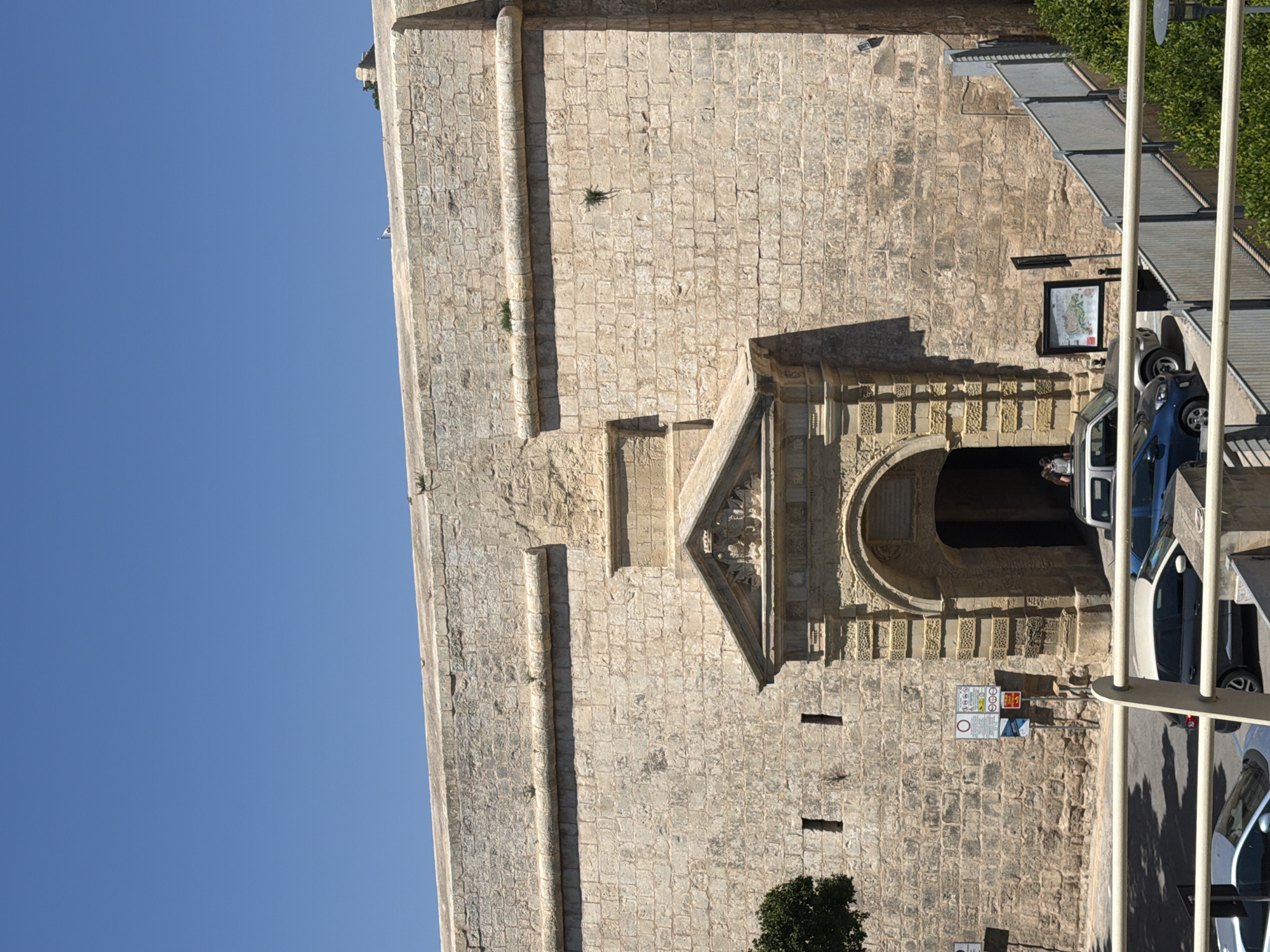 Here is a forensic description of the image:
The image depicts a tall, tan-colored stone building in an urban setting. A clear blue sky fills the upper left portion of the frame. The building's facade features a series of raised bands running vertically along its length. A decorated arched entrance is visible, with a triangular structure above the arch containing intricate carvings.
Adjacent to the building is a pedestrian walkway with metal railings. Several cars are parked on the street beside the building. A person can be seen standing inside the archway. Various signs are attached to the building's wall near the entrance. A small green tree is at the bottom right of the frame. The city of the building is unknown.