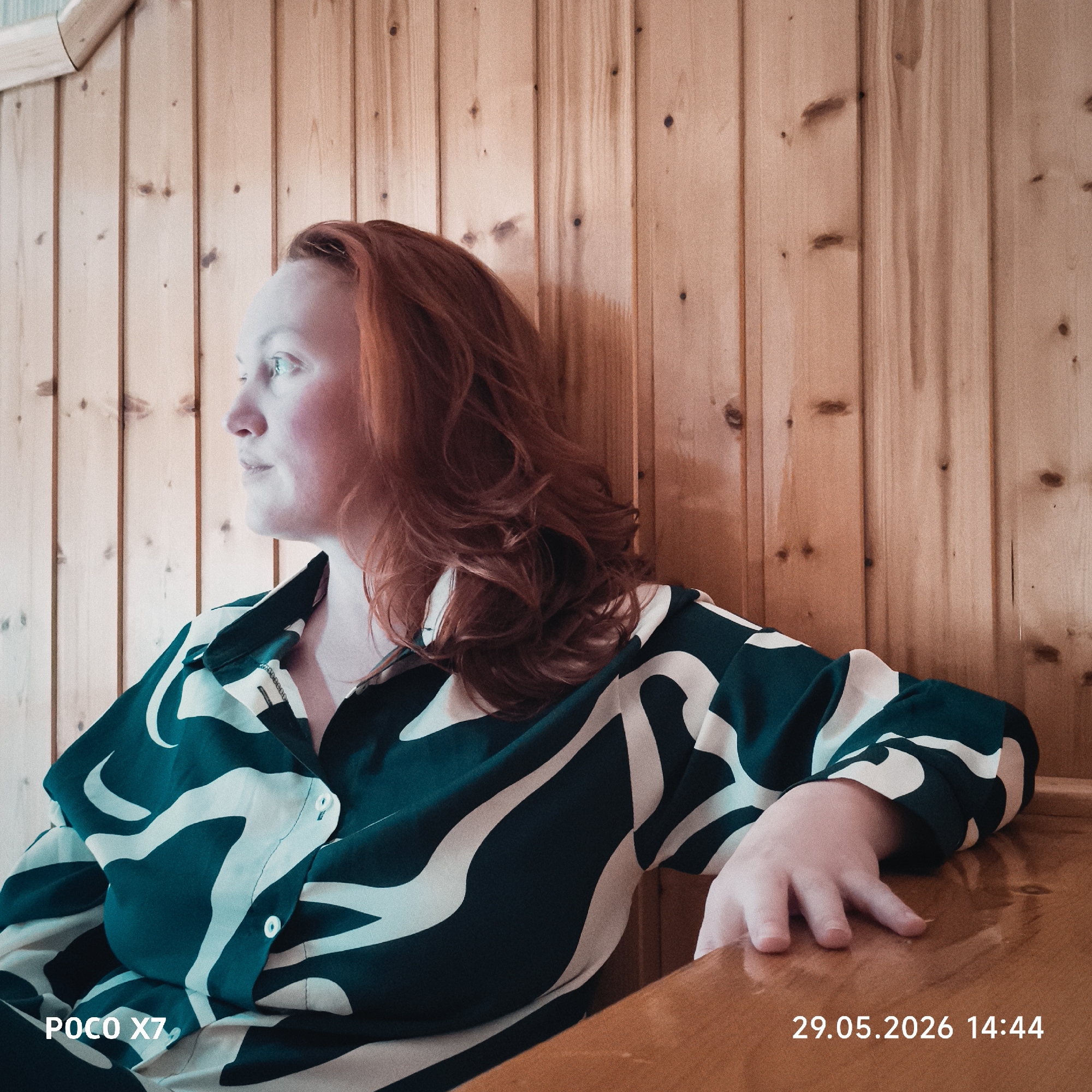 A medium shot depicts a fair-skinned woman with red hair seated against a wooden wall, her gaze directed to the left. She wears a patterned shirt with black and white abstract designs. Her right hand rests on a wooden surface. The wall behind her is composed of light brown vertical wooden planks. In the lower-left corner, "POCO X7" is printed, while "29.05.2026 14:44" is visible in the lower-right corner. The location is Unknown, Unknown.