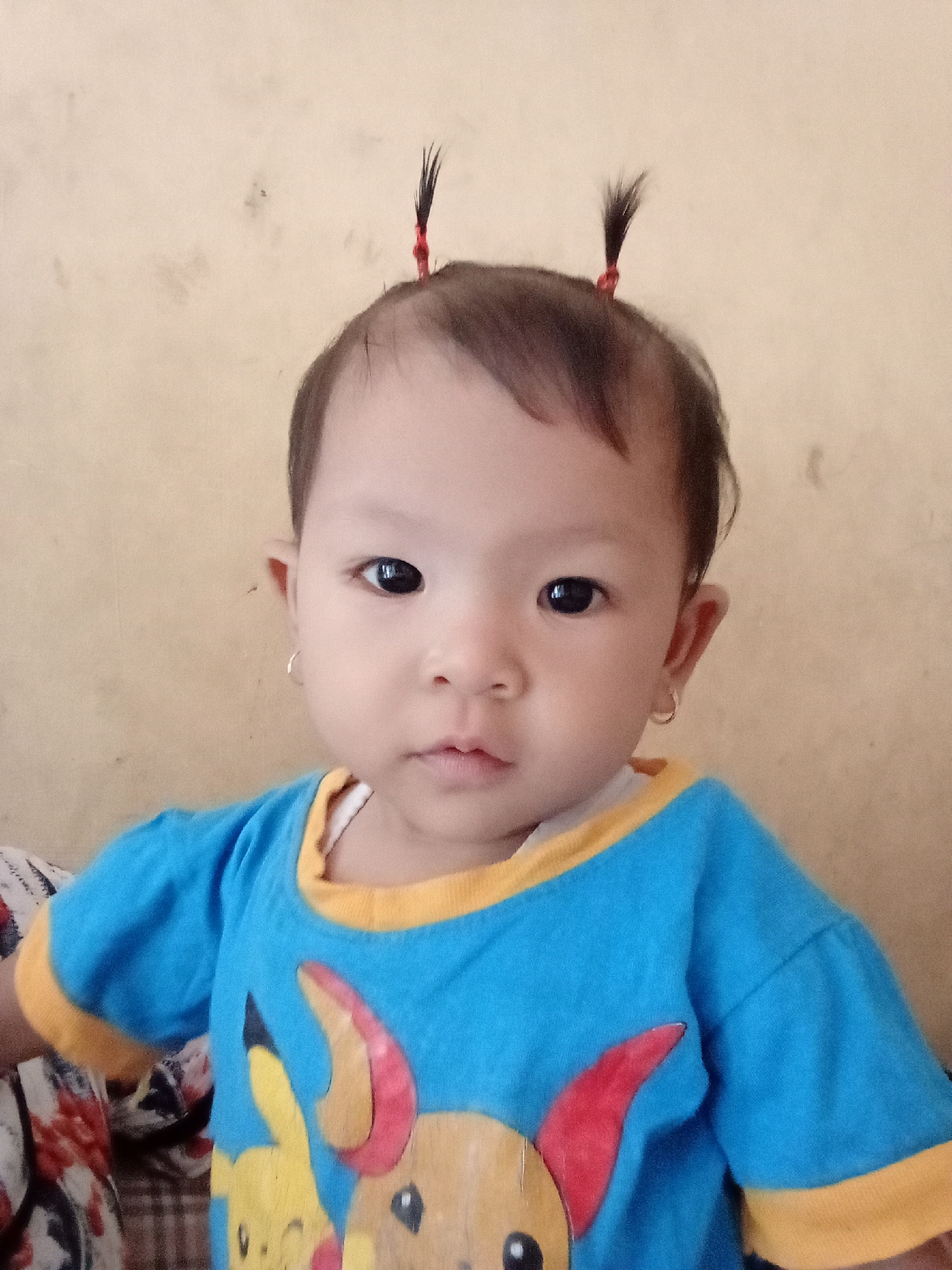 A close-up, head-on shot shows a young child with two small ponytails tied with red string against a plain, beige wall. The child is wearing a blue t-shirt with a yellow collar and cuffs. The t-shirt features a graphic of Pikachu, a popular Pokémon character, on the front. The child has dark hair, dark eyes, and is wearing a small hoop earring in their left ear. The background is out of focus.