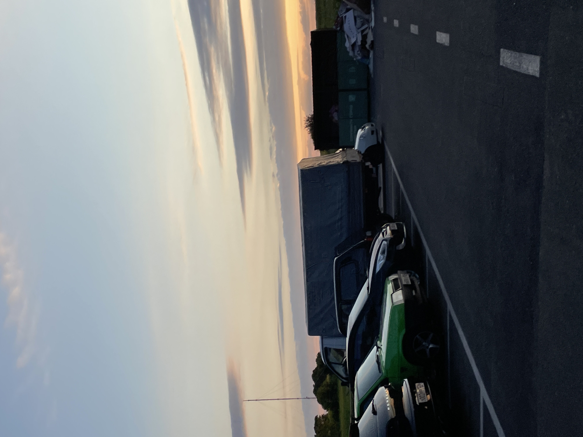 The image depicts an outdoor parking lot at dusk in Stavropol, Russia. In the foreground, several cars are parked, including a black Porsche and a green Lada. The cars are positioned on asphalt with white parking space lines. In the middle ground, a large grey tarp-covered truck is parked, along with other vehicles and large green containers, possibly for waste disposal. The background features a rural landscape with trees and a distant radio tower against a sky with soft, elongated clouds illuminated by the setting sun. The lighting suggests the time is shortly after sunset.