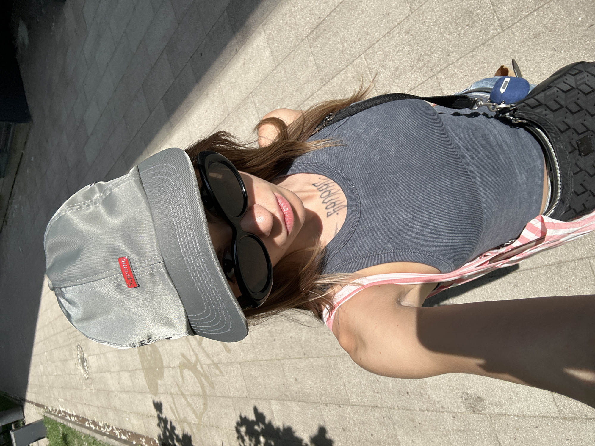 A young woman is taking a selfie on a sunlit, paved sidewalk in Moscow, Russia. She is wearing a grey baseball cap with a red "Supreme" logo, black oval sunglasses, and a grey ribbed tank top. A pink and white striped strap is visible over her right shoulder. She has dark hair and a tattoo on her neck. A black crossbody bag with a patterned design is slung across her body, and a small blue pouch is attached to it. The ground is made of concrete pavers, and there is a patch of green grass to the left. Shadows from trees and possibly a building are cast across the sidewalk. The lighting suggests it is daytime, likely late morning or early afternoon.