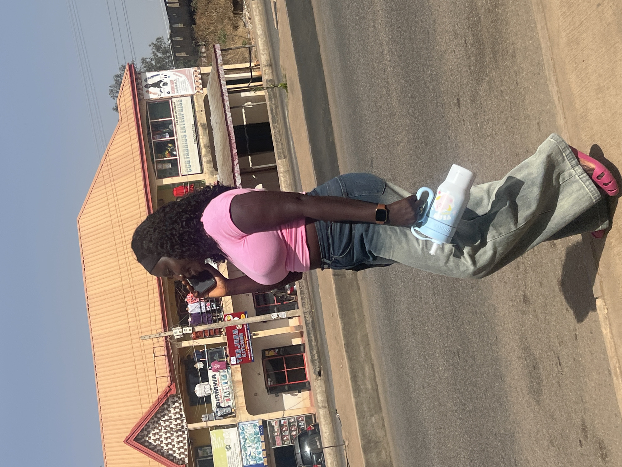 The image displays a woman in profile, walking or standing on a street in bright daylight. She has dark, curly hair and is wearing a pink short-sleeved top, light blue jeans, and pink sandals. A dark smartwatch is visible on her left wrist. She holds a dark smartphone to her left ear, engaged in a call, and carries a light blue and white baby bottle or thermos with a handle in her right hand.
The scene is an outdoor urban or semi-urban environment, with a clear blue sky overhead and strong sunlight casting distinct shadows, suggesting mid-day. A paved road or sidewalk runs parallel to a row of commercial buildings. These buildings feature light-colored walls and reddish-brown roofs, indicative of local architecture.
Several signs are visible on the buildings:
* A large sign clearly reads "C.G.U. FABRICS ENTERPRISE" with "CAC BN 267380" and a phone number "07033709033".
* Above this, another sign displays "FASHIONCITY" and "PLUS WORLD".
* Further down the street, a red sign advertises "TEEJEESS KITCHEN".
* Other signs, partially obscured, show text such as "DISMWA".
The overall impression is a vibrant commercial area in Jos, Nigeria, during a sunny day. The woman appears focused on her phone call while navigating the street.
