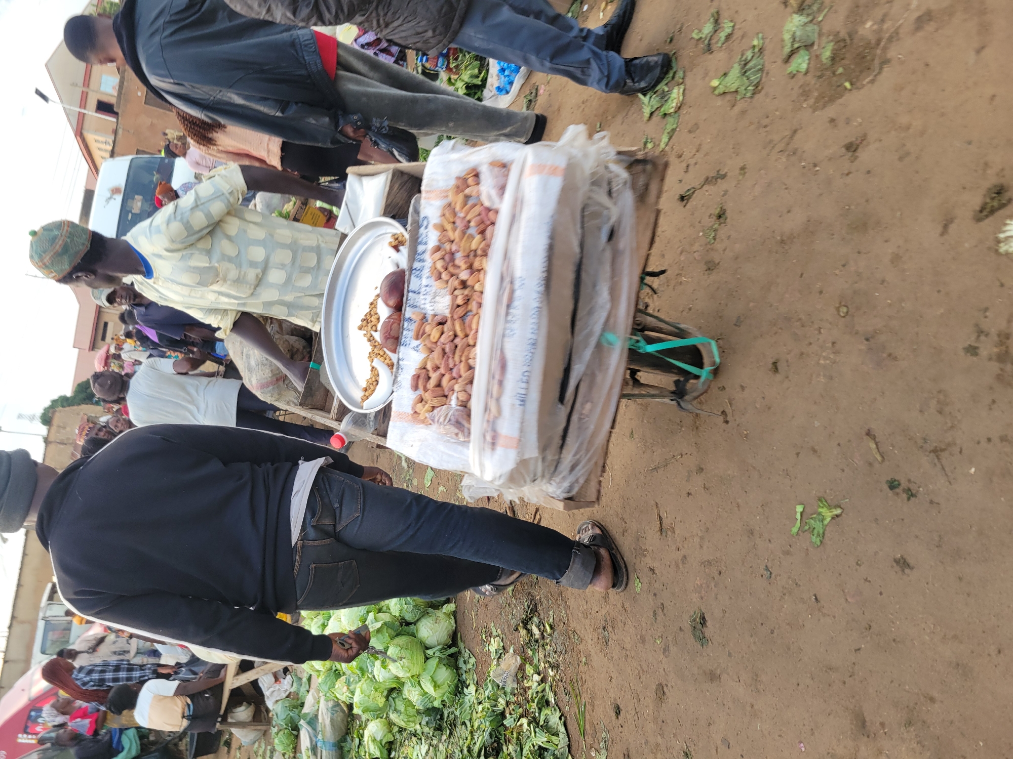 This media file depicts a busy outdoor market scene, likely in Gura Top, Nigeria. Several people are present, including vendors and shoppers. The foreground is dominated by a person wearing a black hooded sweatshirt and dark jeans, with their back to the camera, and they are holding a metal object. To the left, a pile of cabbages is visible on the ground, with some scattered leaves.
Behind the person in the black sweatshirt, a market stall or cart is laden with produce, primarily nuts encased in plastic packaging. A silver tray on the cart holds a selection of these nuts and what appear to be some round, reddish-brown items, possibly fruits or vegetables.
Further back, other individuals are seen browsing or working at the market. One person, wearing a light-colored patterned shirt, is looking towards the market stall. Another person in a black jacket is walking away. A white van is parked in the background, suggesting transportation is readily available. The ground appears to be dirt or sand, common in open-air markets.
The overall impression is of a lively marketplace. The lighting suggests it is daytime, possibly late morning or afternoon, with no strong indications of specific weather conditions beyond the outdoor setting. The presence of diverse people and goods points to a functioning local economy. No discernible text is clearly legible in the image.