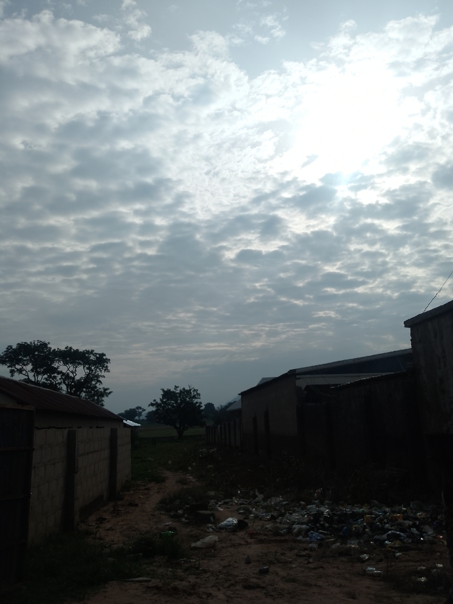 The image depicts an overcast sky with the sun faintly visible through the clouds. In the foreground, there is a dirt path littered with a large amount of trash, including plastic bottles and bags. To the left of the path is a small building constructed from cinder blocks with a corrugated metal roof. Further back, more buildings and fences are visible. On the left side of the image, a large tree with dense foliage stands out. The scene appears to be an outdoor area, likely a residential or informal settlement in the city of Serti, Nigeria, given the presence of rudimentary structures and visible refuse. The lighting suggests it is daytime.