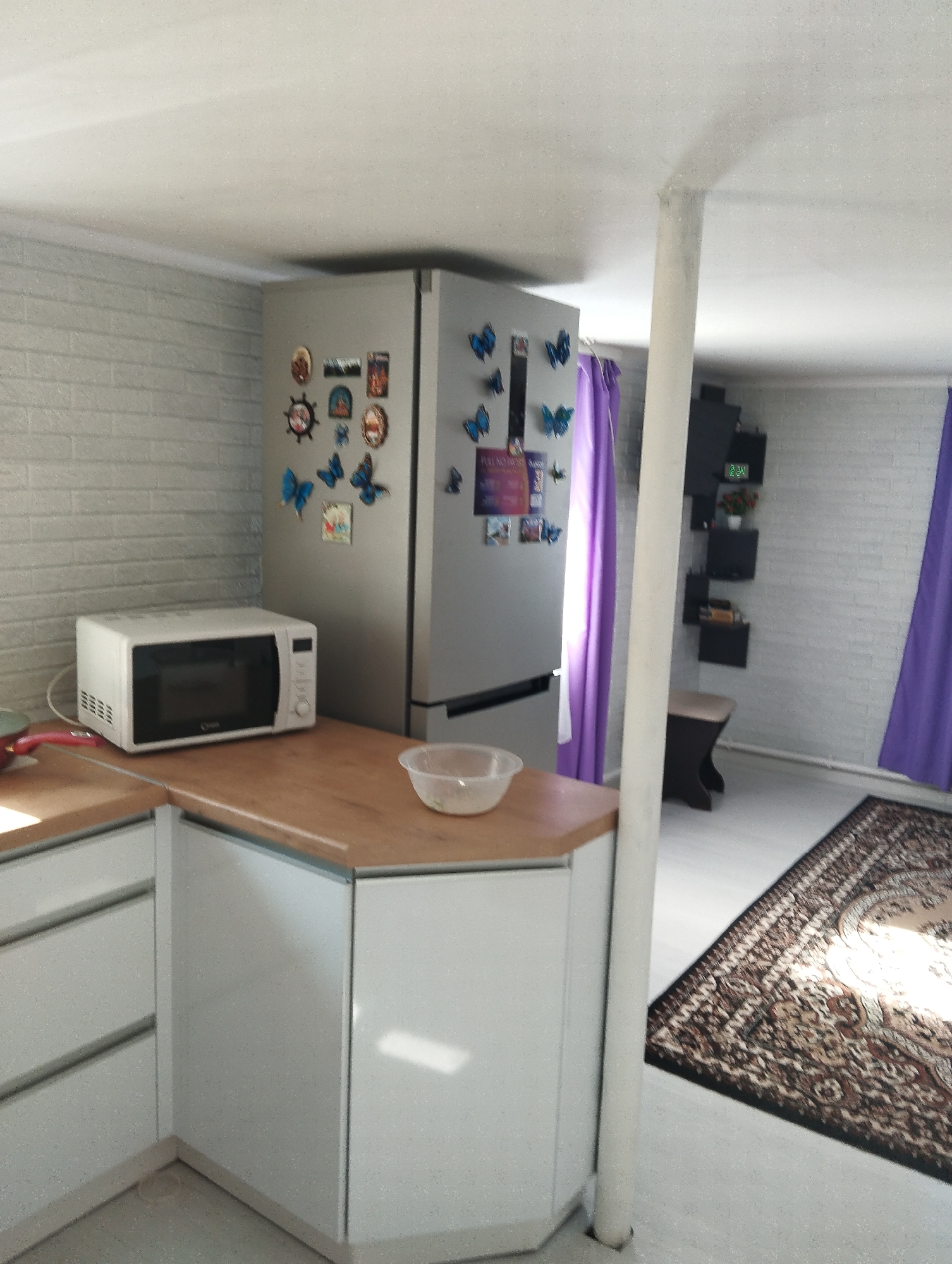 The image depicts an interior space in Krasnogorskoye, Russia. A kitchen area with light grey brick walls is visible on the left, featuring white cabinets with light wood countertops, a white microwave, and a stainless steel refrigerator adorned with magnets. A clear plastic bowl sits on the counter. A white structural pole is positioned near the kitchen area. Beyond the pole is a room with a black shelving unit on the wall, a small stool, purple curtains, and a brown patterned rug on the floor. The lighting is bright, suggesting daylight.
