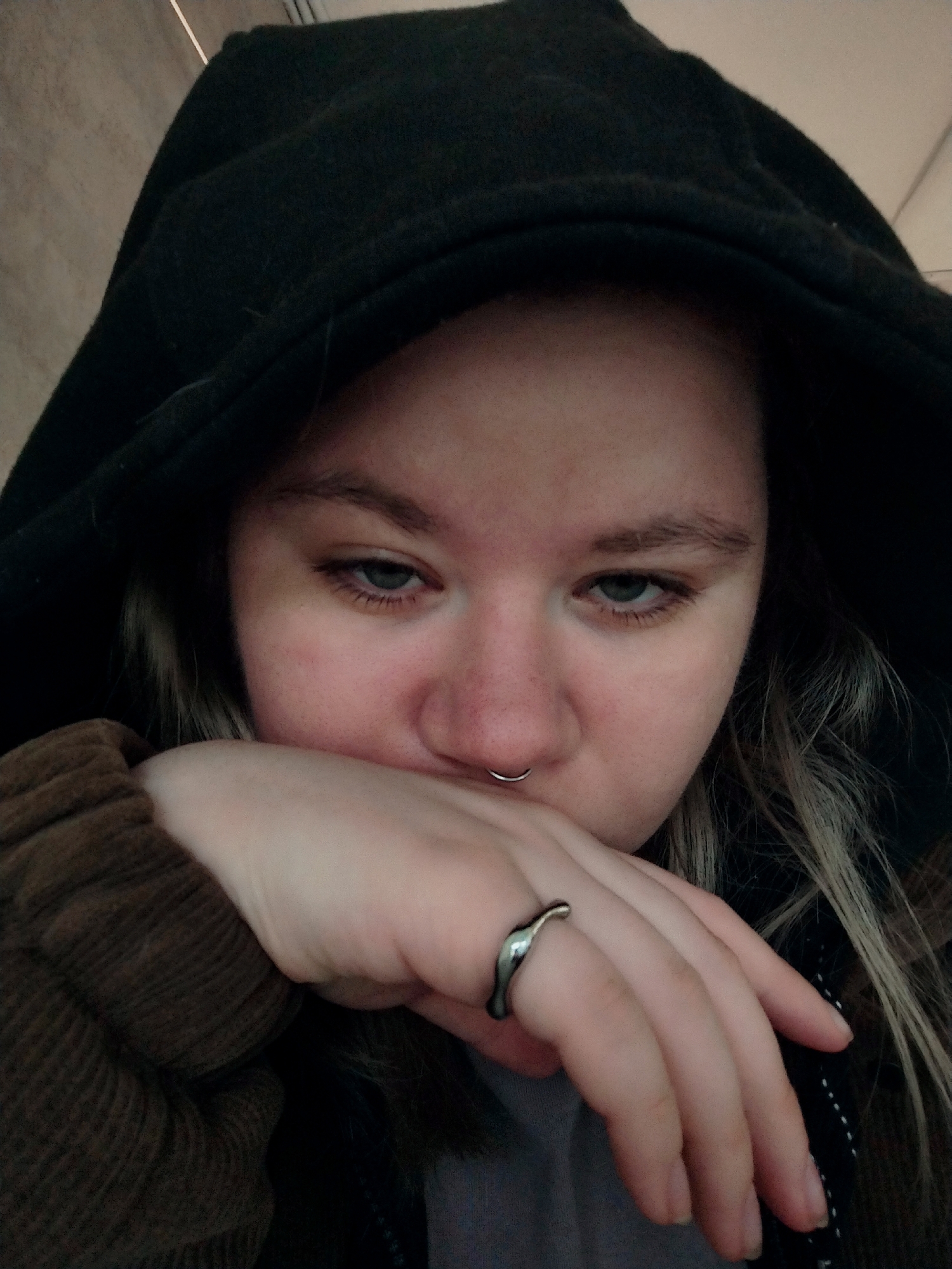 A young, fair-skinned person with light brown hair is shown from the shoulders up, looking downwards with a neutral expression. They are wearing a black hooded sweatshirt, a brown corduroy jacket, and a silver ring with an abstract, wavy design on their left index finger. A silver hoop is visible in their septum. The background is out of focus and appears to be an indoor, possibly urban, environment with neutral-colored walls. The lighting is soft and diffused.