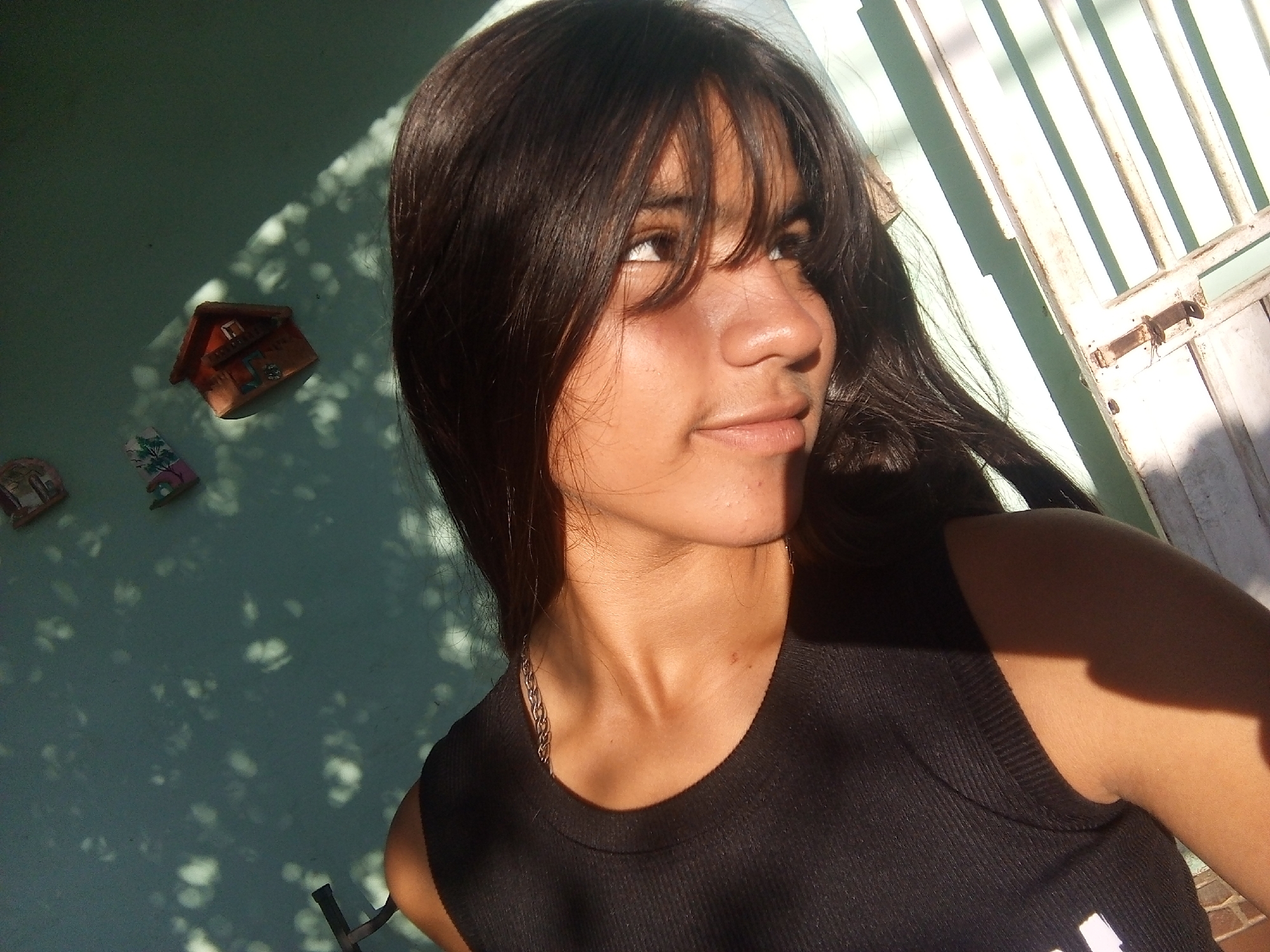 A young woman with long, dark hair and bangs is positioned in the foreground, wearing a black tank top and a silver chain necklace. She is looking upwards and to the left of the frame. Behind her is a turquoise wall with decorative ceramic birdhouses and shadow patterns cast upon it. To the right of the woman is a white metal gate with vertical bars. The setting appears to be outdoors. No specific location can be identified in Juan Griego, Venezuela.