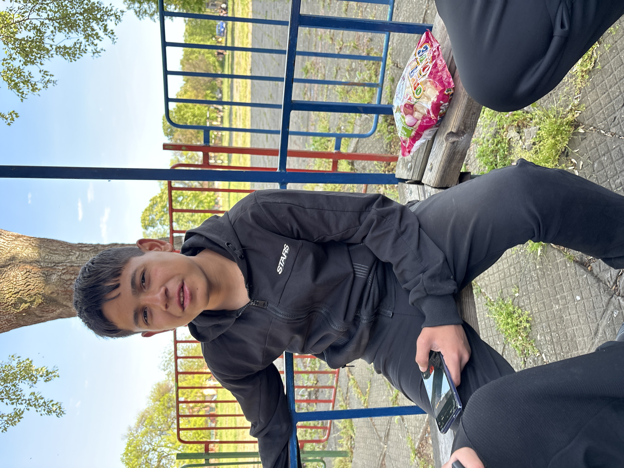 The image depicts an outdoor scene in Glava, Bulgaria, likely a park or playground, on a bright, sunny day with a clear blue sky. The primary subject is a young person, possibly male, with short dark hair, who is sitting on a wooden bench and looking directly at the camera with a slight smile, conveying a relaxed demeanor. They are dressed in a black zipped hoodie with the word "STARS" printed in white on the left chest, and matching black sweatpants.
Two other individuals are partially visible. To the right of the main subject, another person is seated on the same bench, with only their black-clad leg and part of their torso visible. Next to them, resting on the bench, is a colorful snack bag, possibly containing chips or nuts, with Cyrillic text partially readable as "БЯЛА," "САЛА," and "ЦАРСКА," which likely refers to a snack brand such as "Белла Царска салата." In the lower right foreground, the legs of a third person are visible, also wearing black pants, holding a smartphone.
The background features prominent blue and red metal railings, suggesting playground equipment or a fence. A large tree trunk is visible on the far left. The ground is a mixture of green grass, scattered vegetation, and a light-colored paved surface. The time of day appears to be midday or afternoon due to the strong sunlight and clear sky, suggesting pleasant weather for outdoor relaxation.