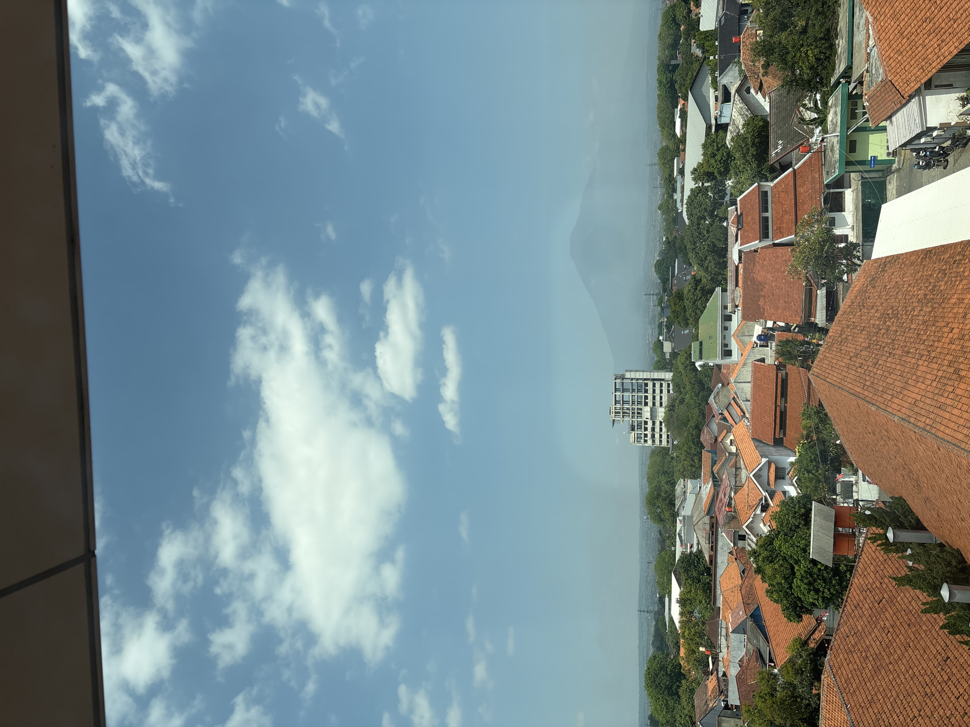 This is a high-angle, daylight, exterior shot of a residential area in Cirebon, Indonesia. The foreground is dominated by the orange-tiled roofs of numerous houses, tightly packed together. Beyond the houses, lush green trees are interspersed with more buildings, including a multi-story white building that appears to be under construction or renovation. In the distance, a hazy mountain range is visible under a bright blue sky with scattered white clouds. The overall scene depicts a dense urban or suburban neighborhood.