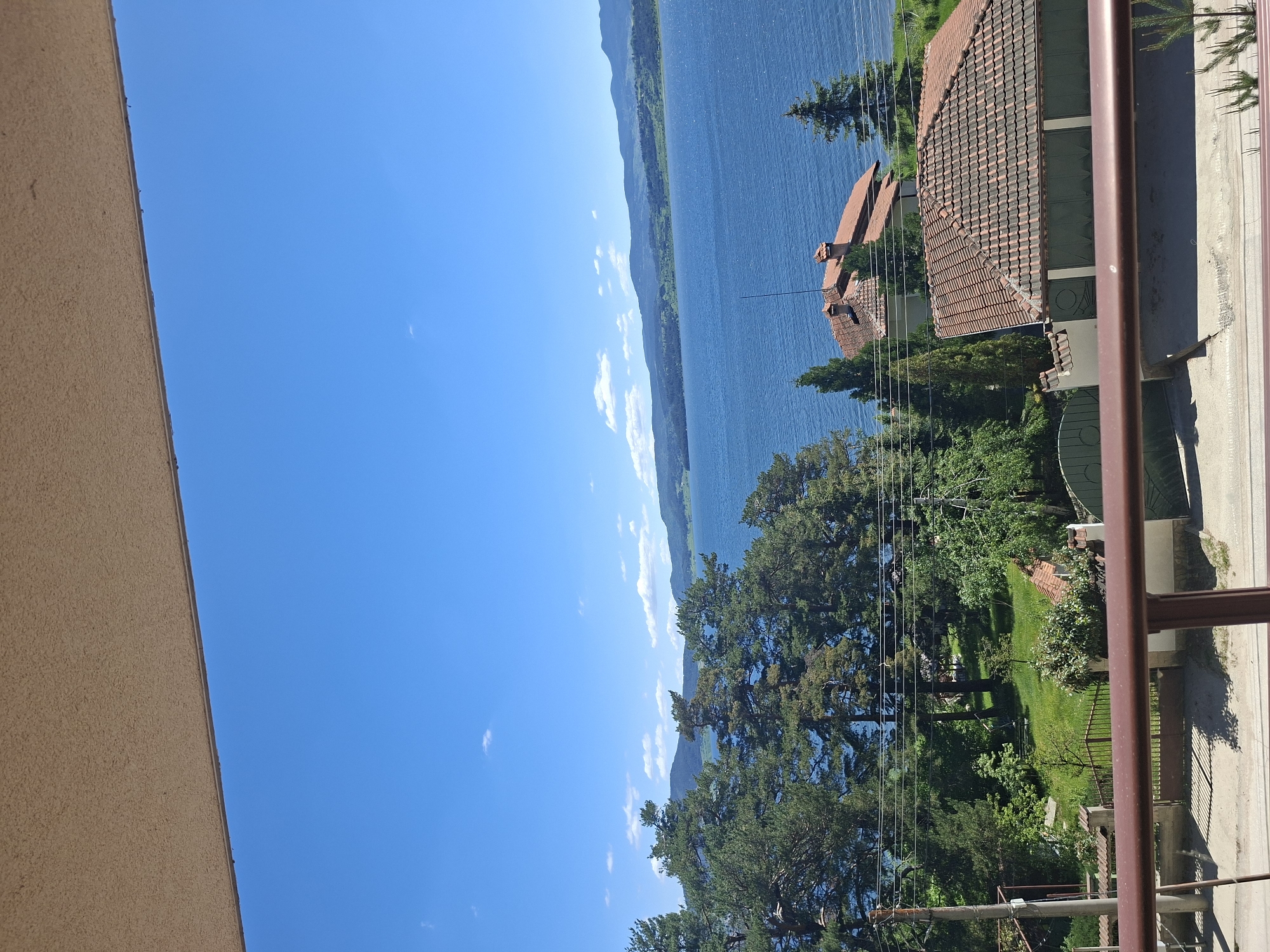 The image presents a scenic view of a lake, with buildings and trees interspersed along the shore. The foreground includes architectural elements of a structure, possibly a balcony, with a brown railing. The buildings in view have red-tiled roofs, suggesting a residential area. Lush greenery is evident, indicating a well-vegetated environment. The lake is the prominent feature, extending to the horizon where it meets the sky. The sky is clear with a light blue hue. The image is taken from an elevated viewpoint, likely from inside a building.